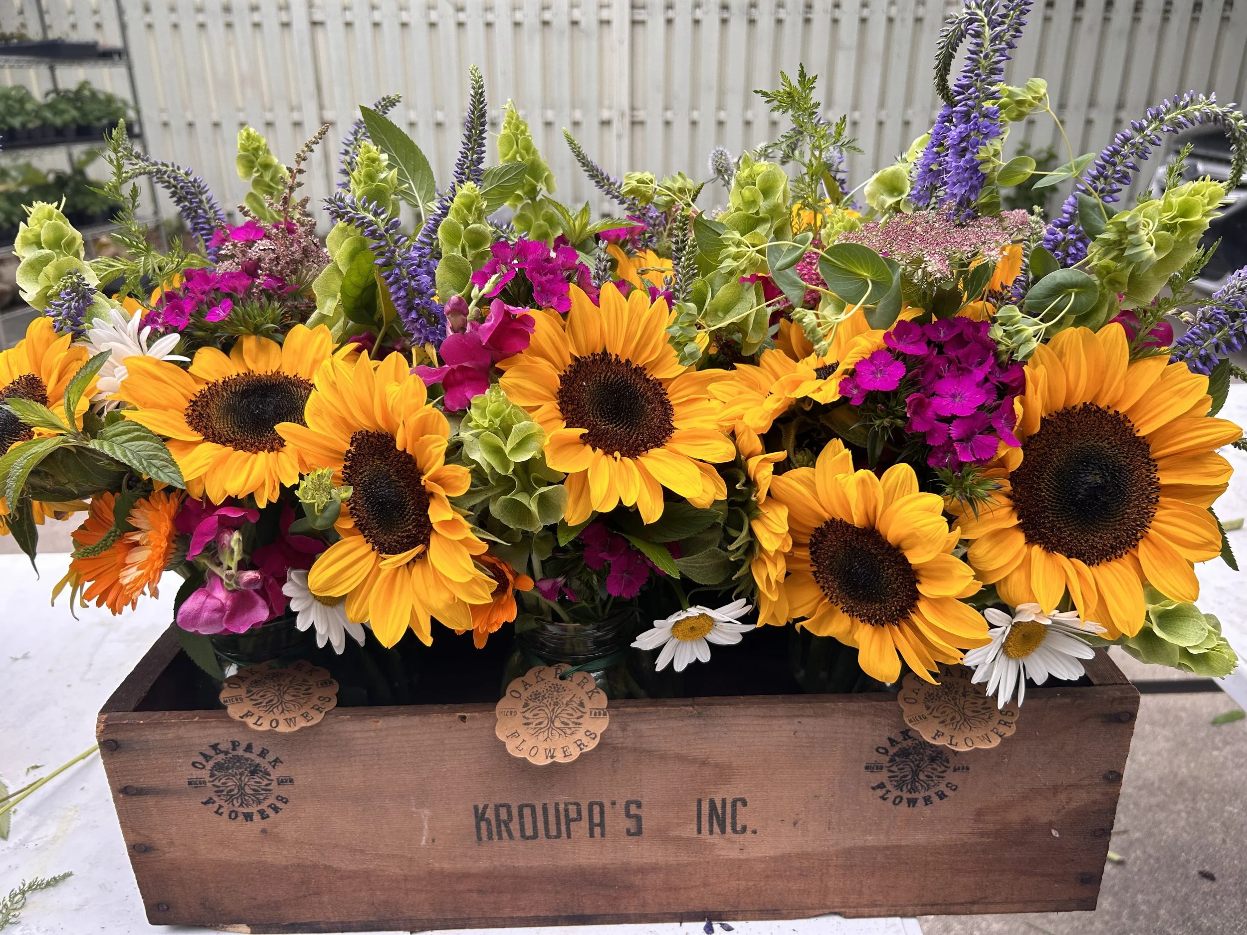 Colorful bouquet of sunflowers, pink, purple, yellow, white, and green flowers in a wooden crate labeled 'Kroupa's Inc.' with additional branding markings.
