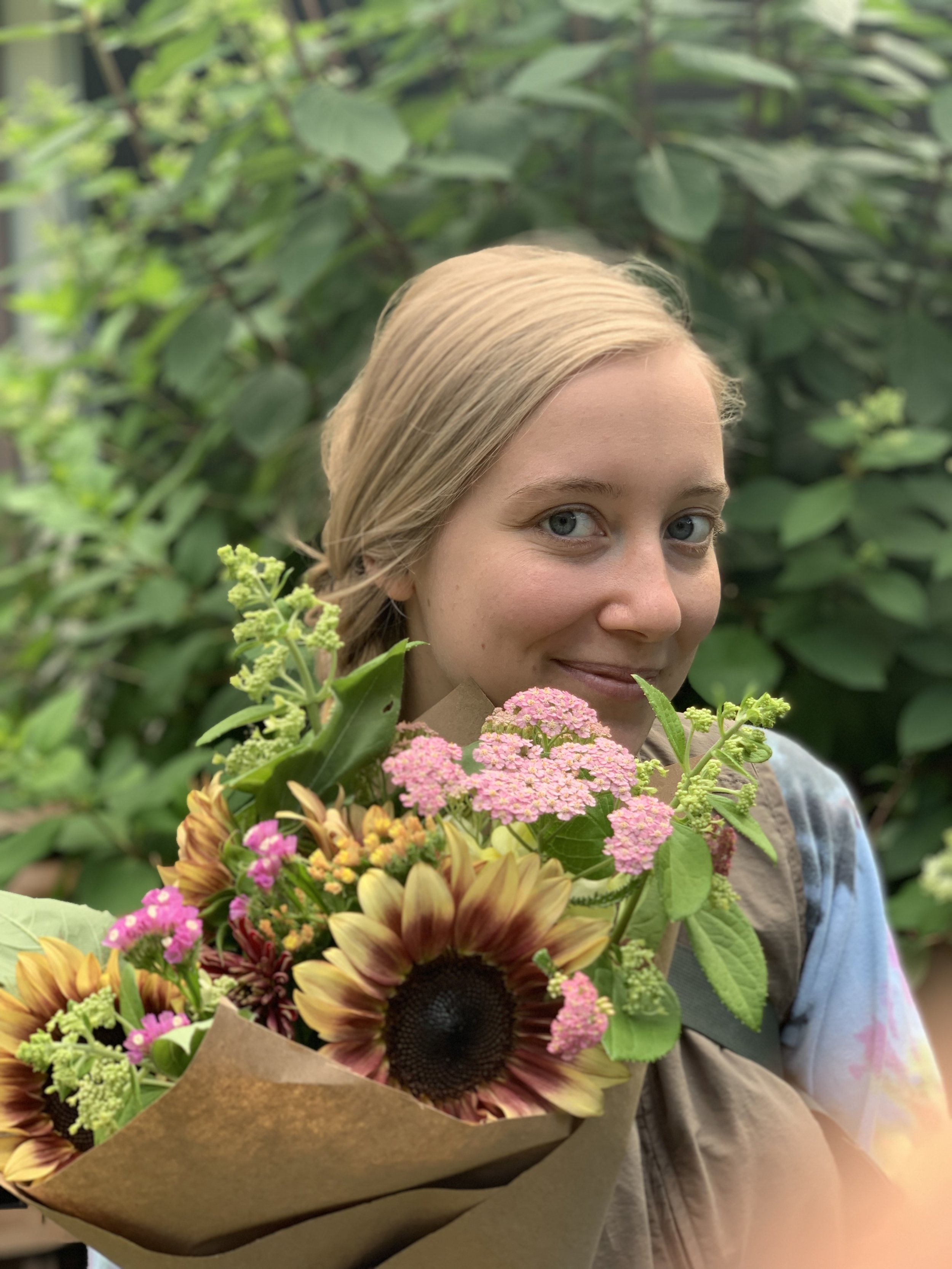 A young woman with blonde hair holding a bouquet of colorful flowers, including sunflowers and pink blossoms, outdoors with green foliage in the background.