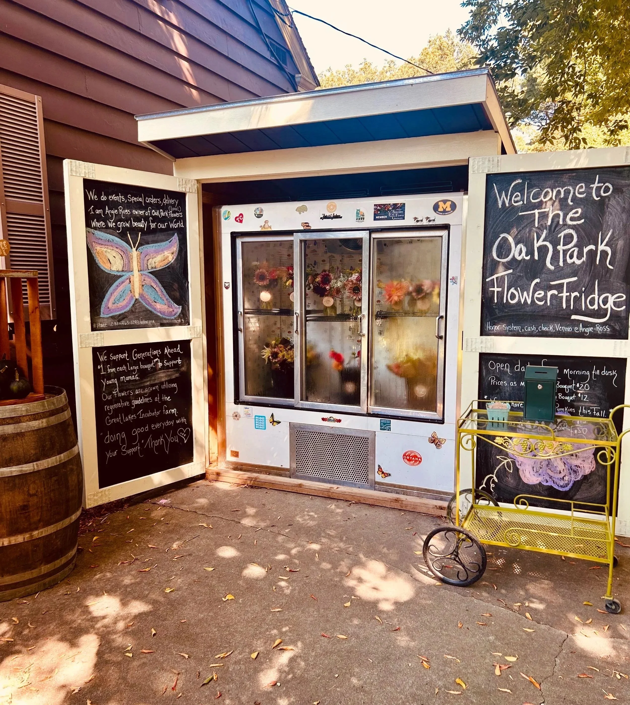 A flower fridge with handwritten blackboards on each side. The right blackboard welcomes visitors to The OakPark Flower Fridge and lists payment options. The left blackboard has a butterfly drawing and information about flower arrangements and support for local farmers. The fridge contains various bouquets of colorful flowers visible through the glass doors. There is a small yellow cart with decorative details in front of the right blackboard and some fallen leaves on the ground.