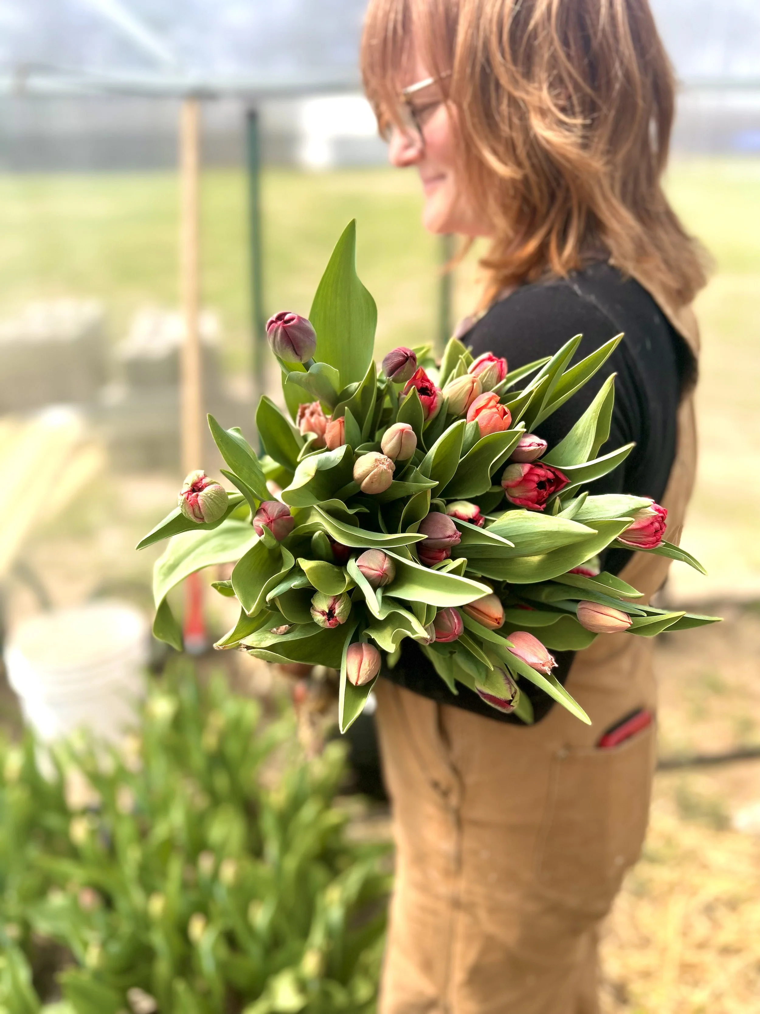A woman with red hair and glasses holding a bouquet of pink and white tulips inside a greenhouse.