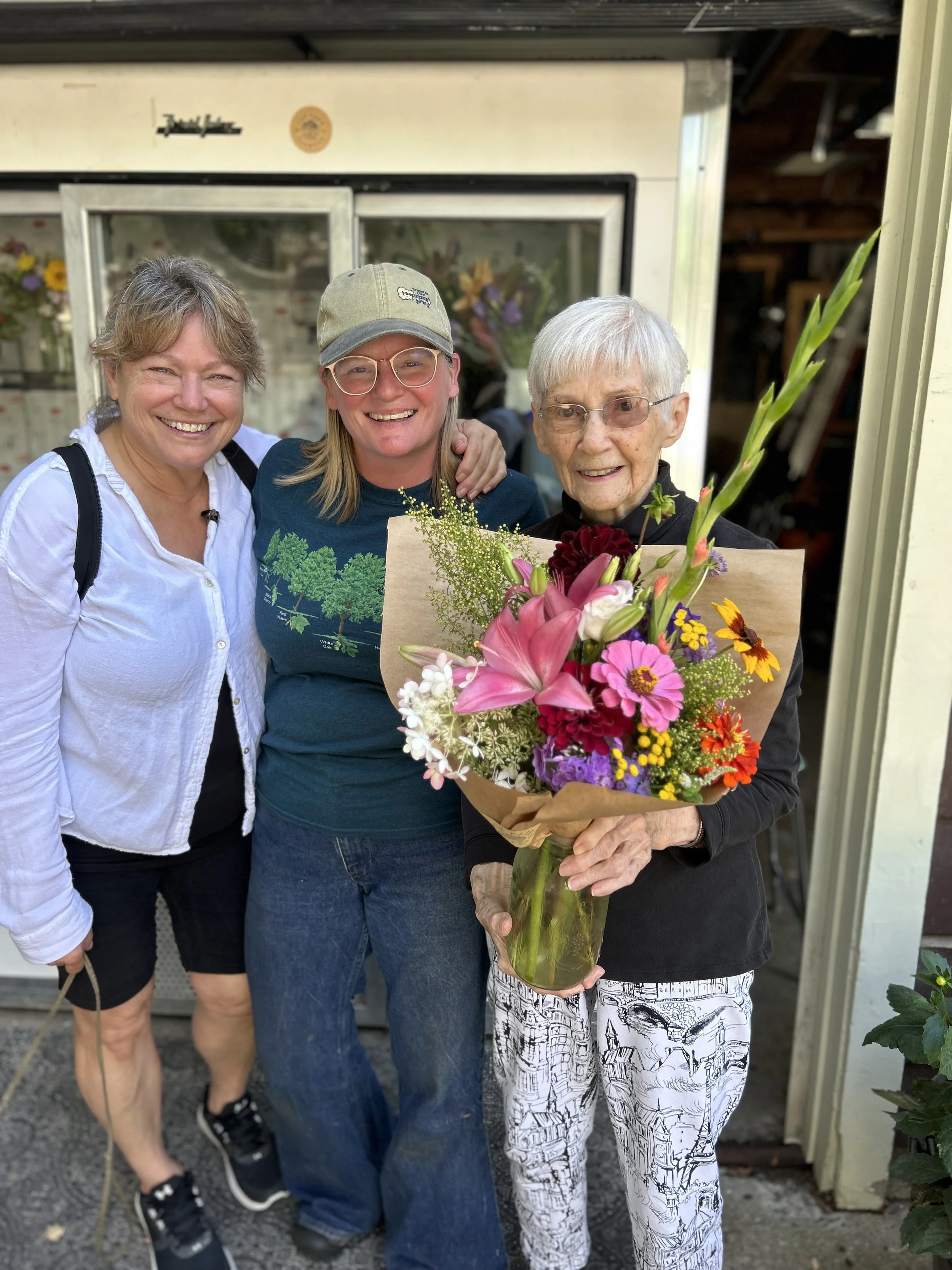 Three women standing together, smiling, with one woman holding a bouquet of colorful flowers.