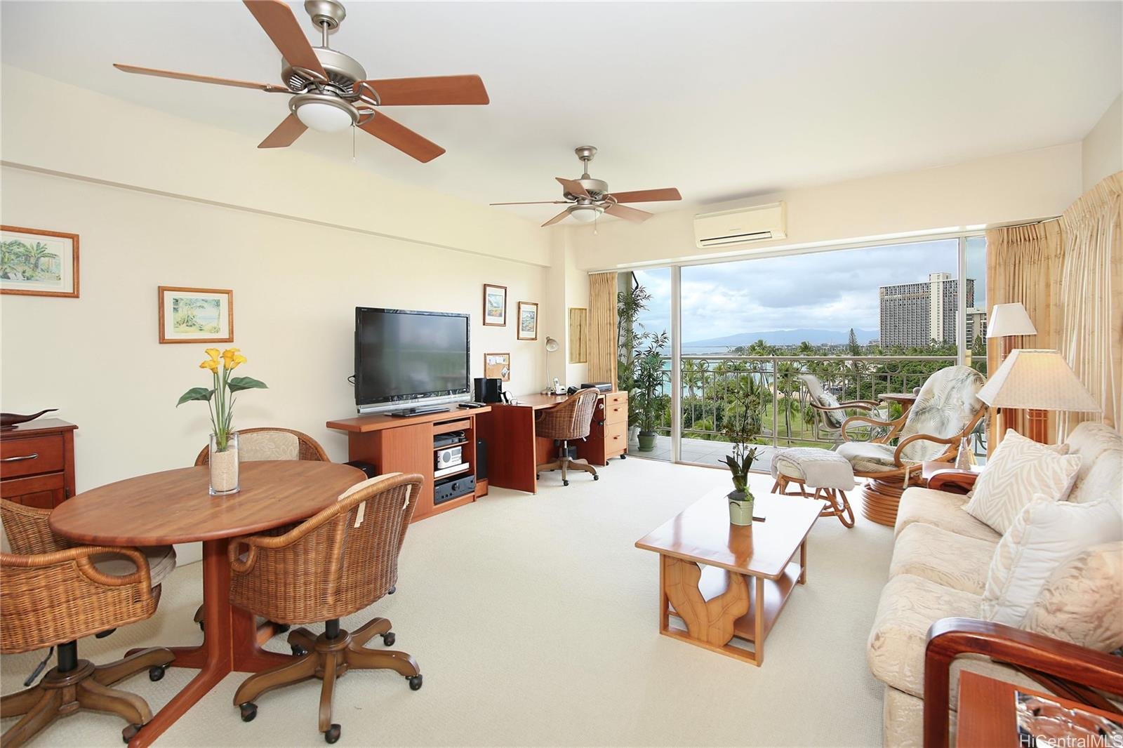 Living room with a view, featuring a white sofa, beige armchair, glass sliding door balcony with outdoor chairs, wooden coffee table, dining table with wicker chairs, ceiling fans, TV, and potted plants.