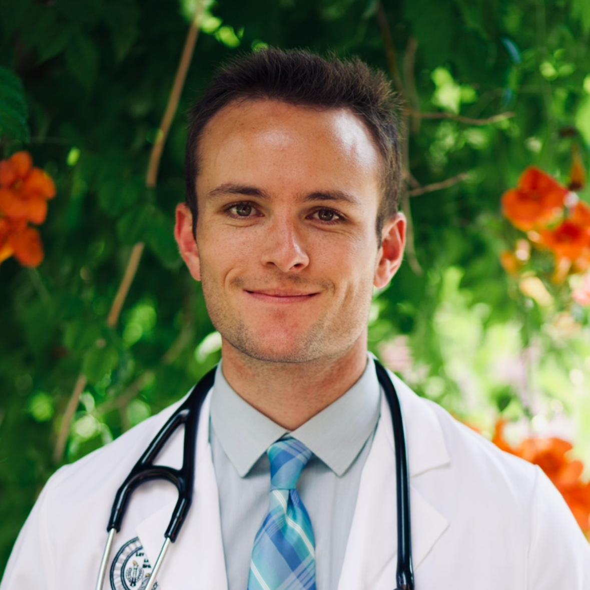 Young male doctor in white coat with stethoscope around neck smiling outdoors with green foliage and orange flowers in the background.