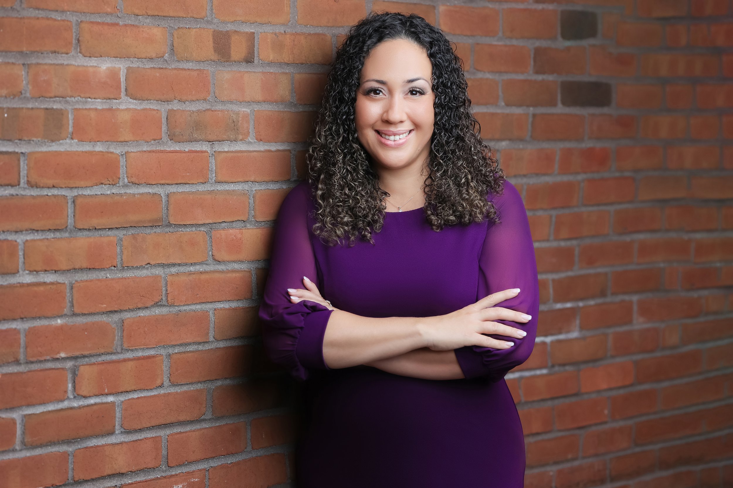 A young woman with curly dark hair wearing a purple dress, smiling, and standing against a brick wall with her arms crossed.