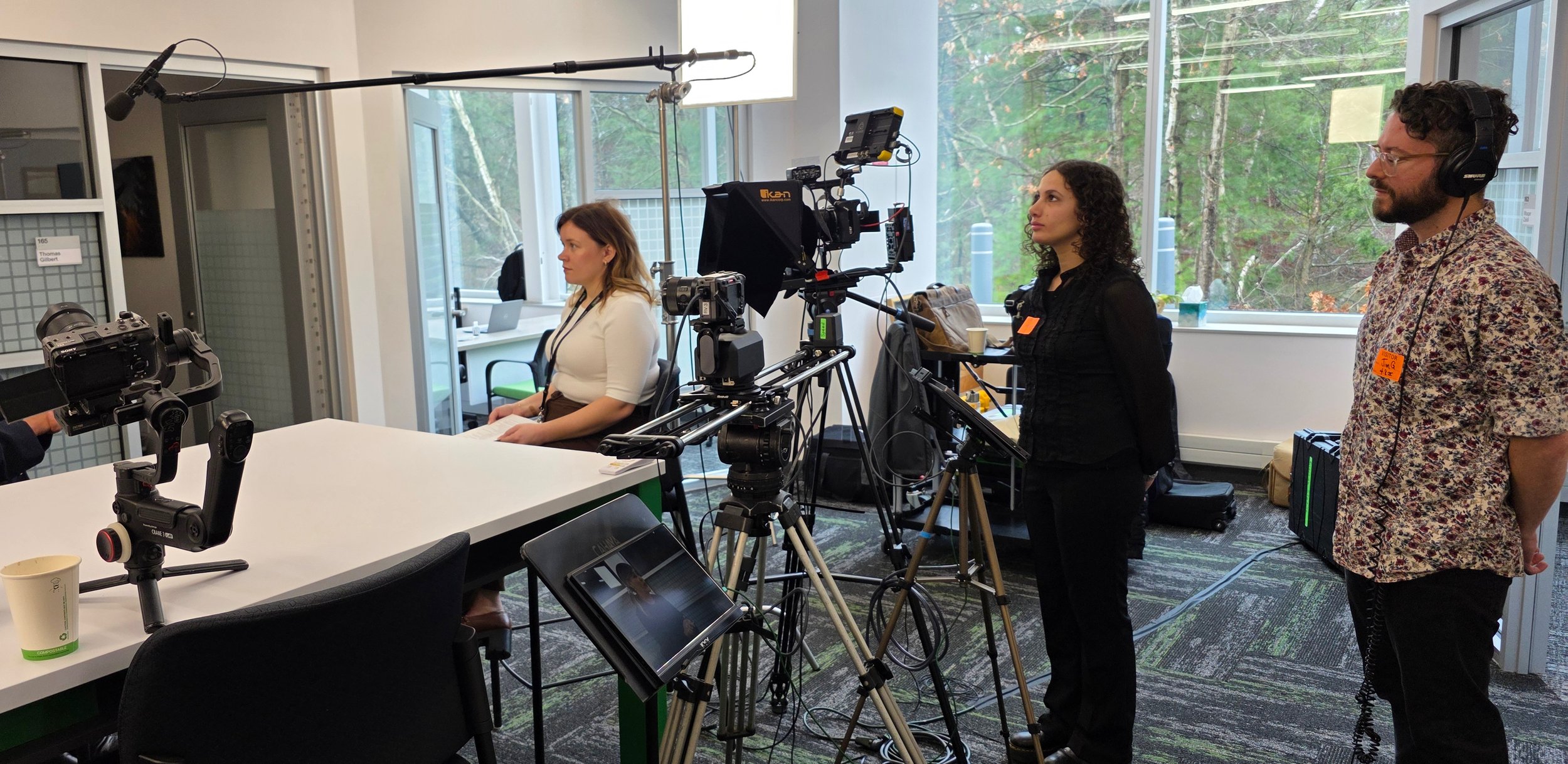 A woman sits at a white table during a video recording, with professional camera and audio equipment aimed at her. Two crew members stand nearby, one with headphones and the other with a patterned shirt, overseeing the shoot in a room with large wind