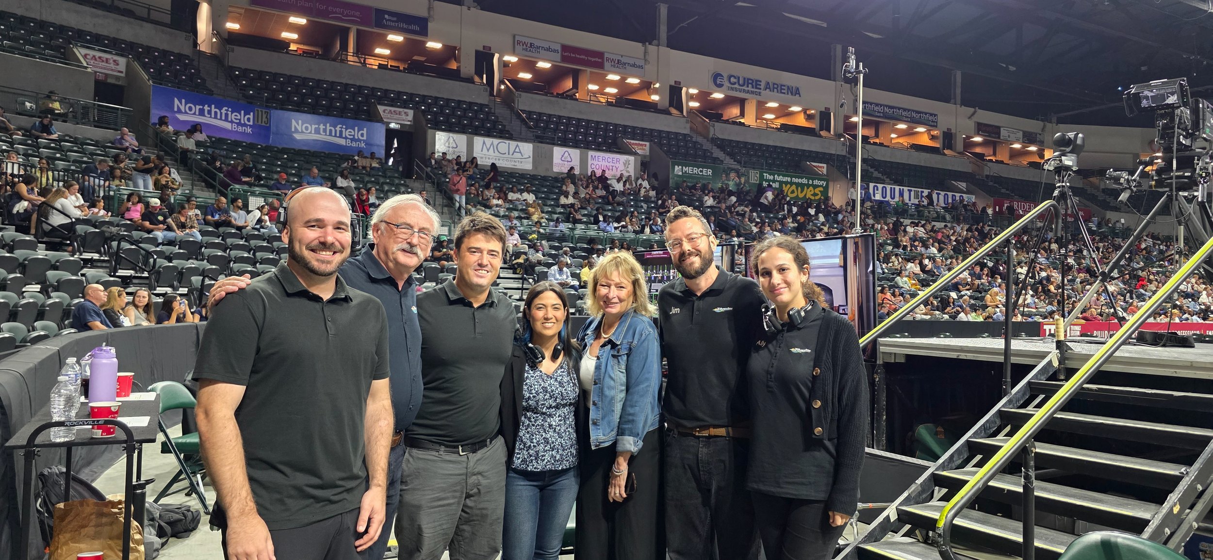 Group of seven people standing on a stage at a sports arena, with seating and audience behind them, and camera equipment to the right.