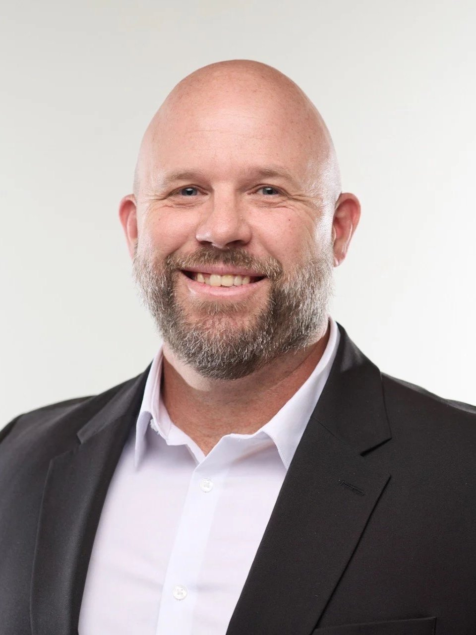 A smiling man with a beard and bald head, wearing a black suit jacket and white shirt, against a plain light background.