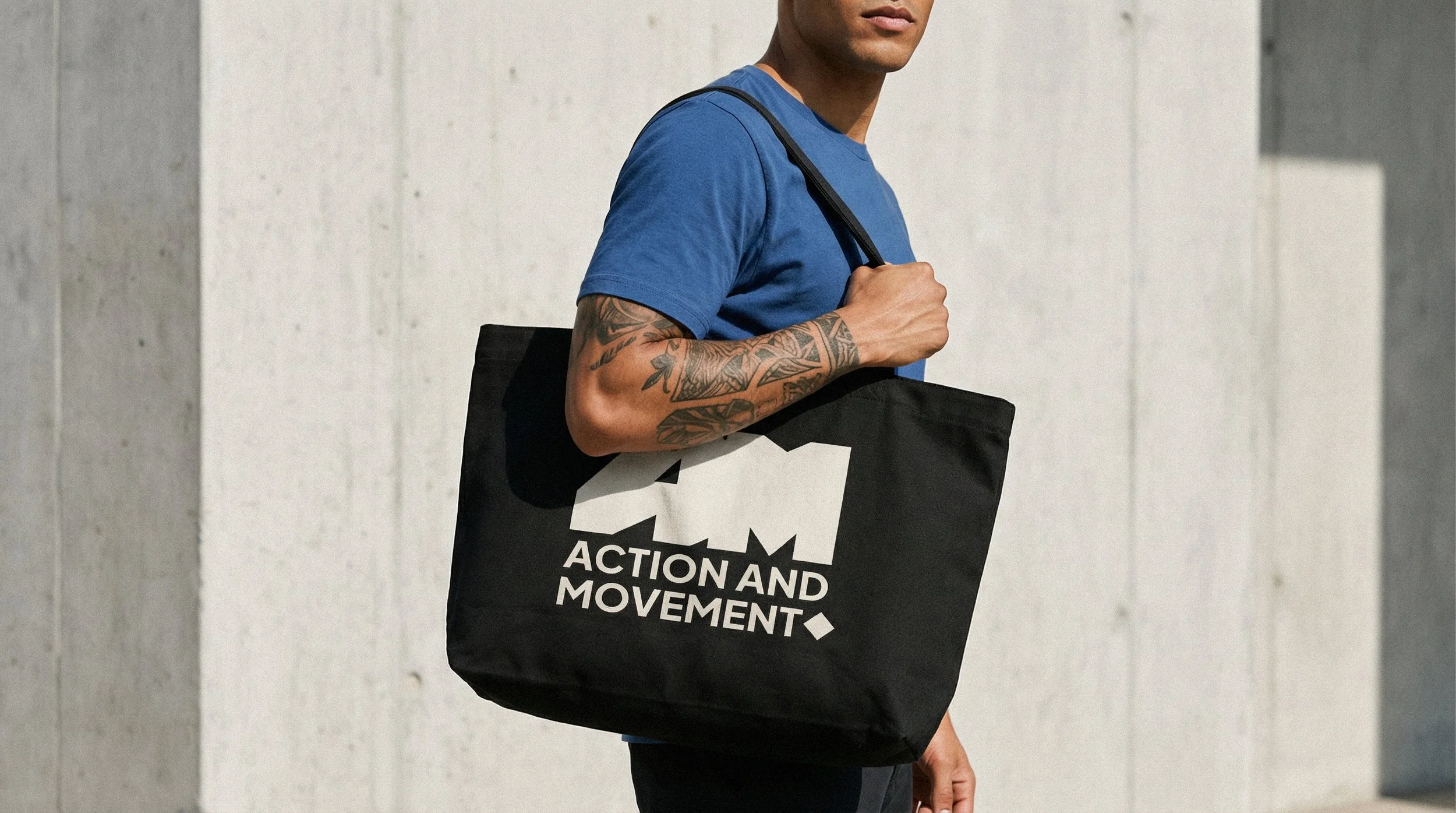 Homem com tatuagens no braço, usando uma camiseta azul, segurando uma bolsa preta com a frase "Action and Movement" escrita em branco, contra um fundo de muro cinza.