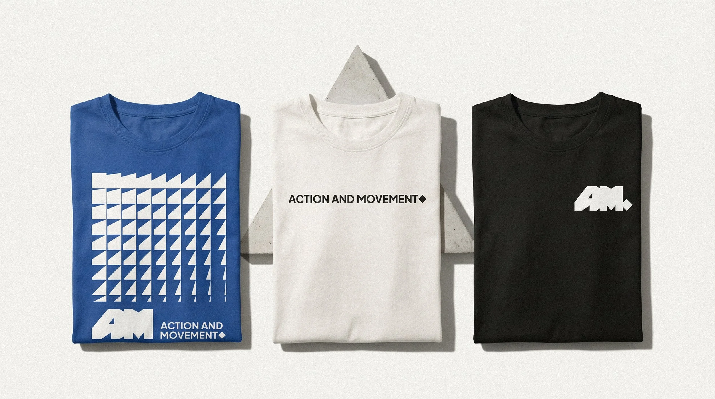 Três camisetas dobradas, uma azul com padrão de triângulos brancos e logo na parte inferior, uma branca com a frase "ACTION AND MOVEMENT" em preto, e uma preta com o símbolo "A.М." em branco, dispostas sobre fundos brancos com elementos geométricos.
