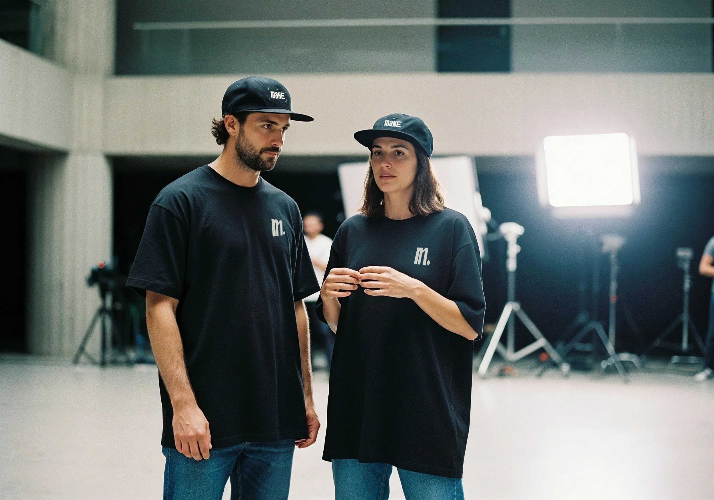 Homens e mulher vestindo camisetas e bonés pretos com logotipo, em ambiente de estúdio com iluminação profissional e equipamentos de filmagem ao fundo.