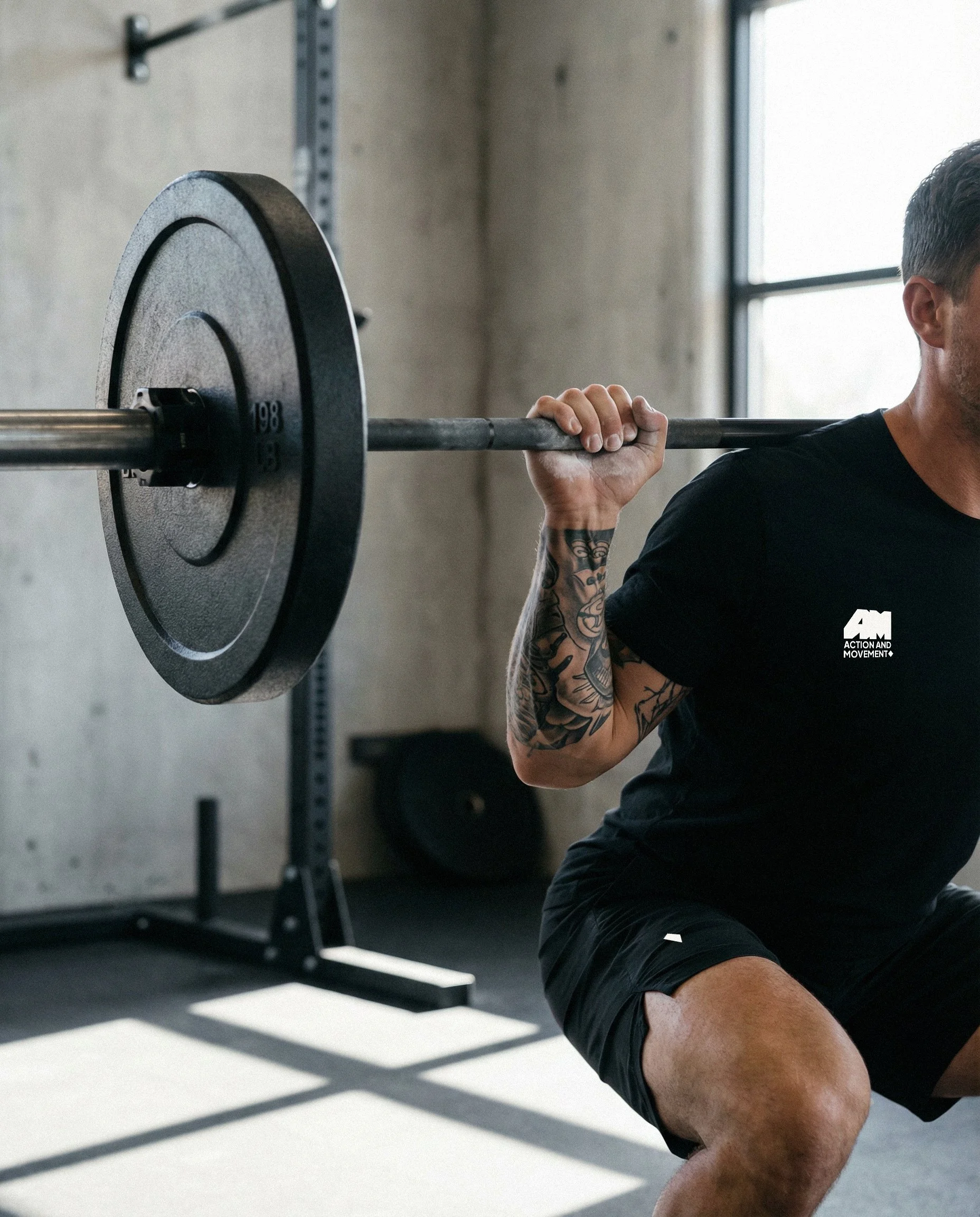 Homem fazendo agachamento com barra no ginásio, de lado, com tatuagens nos braços, usando camiseta preta e shorts.