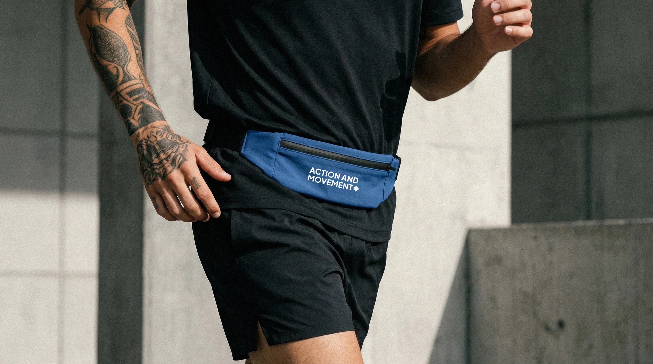 Pessoa correndo com shorts pretos, camiseta preta e pochete azul com a frase 'ACTION AND MOVEMENT' em um ambiente urbano.