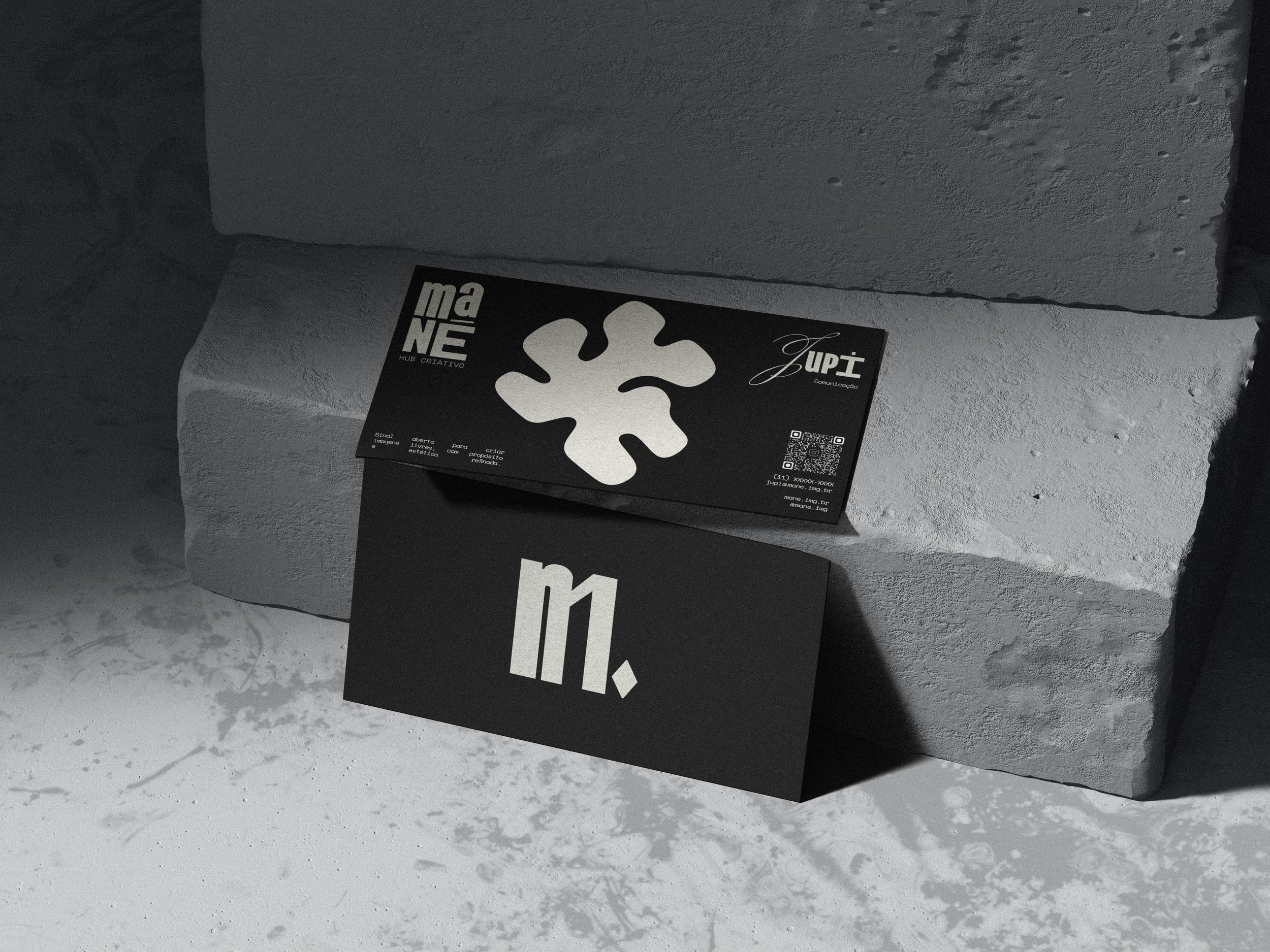 Duas embalagens pretas com caracteres brancos, uma com um símbolo abstrato e outra com uma letra 'm' e um ponto, apoiadas em uma parede de tijolos brancos.