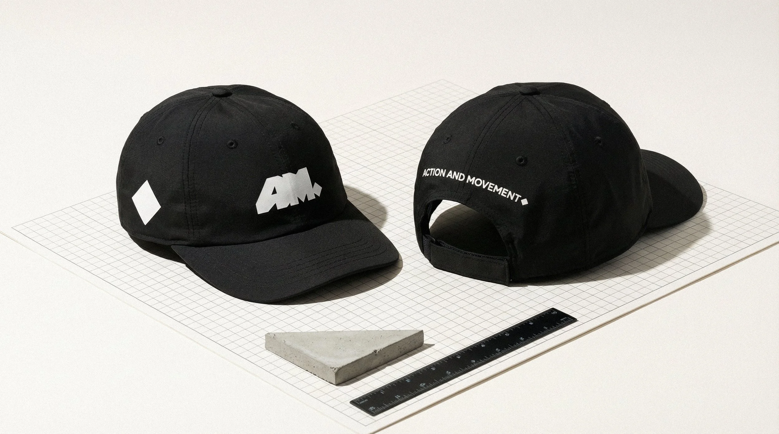 Dois bonés pretos com logotipo e texto, um com logotipo e o outro com frases, ambos posicionados sobre uma superfície com uma régua e um pedaço de material triangular ao lado.