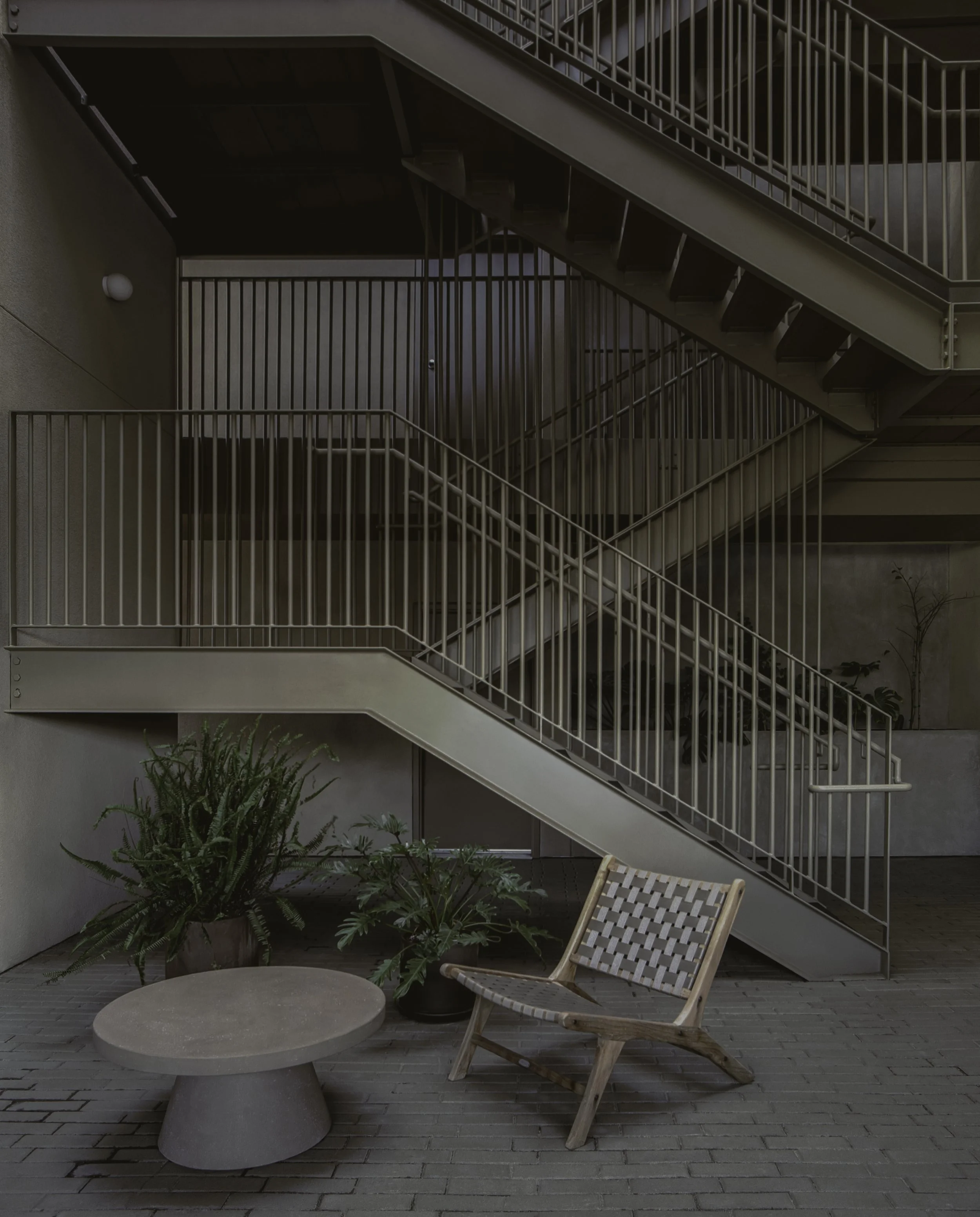 Interior view of a modern building with a staircase with metal railings, potted plants, a round concrete table, and a woven wooden chair.