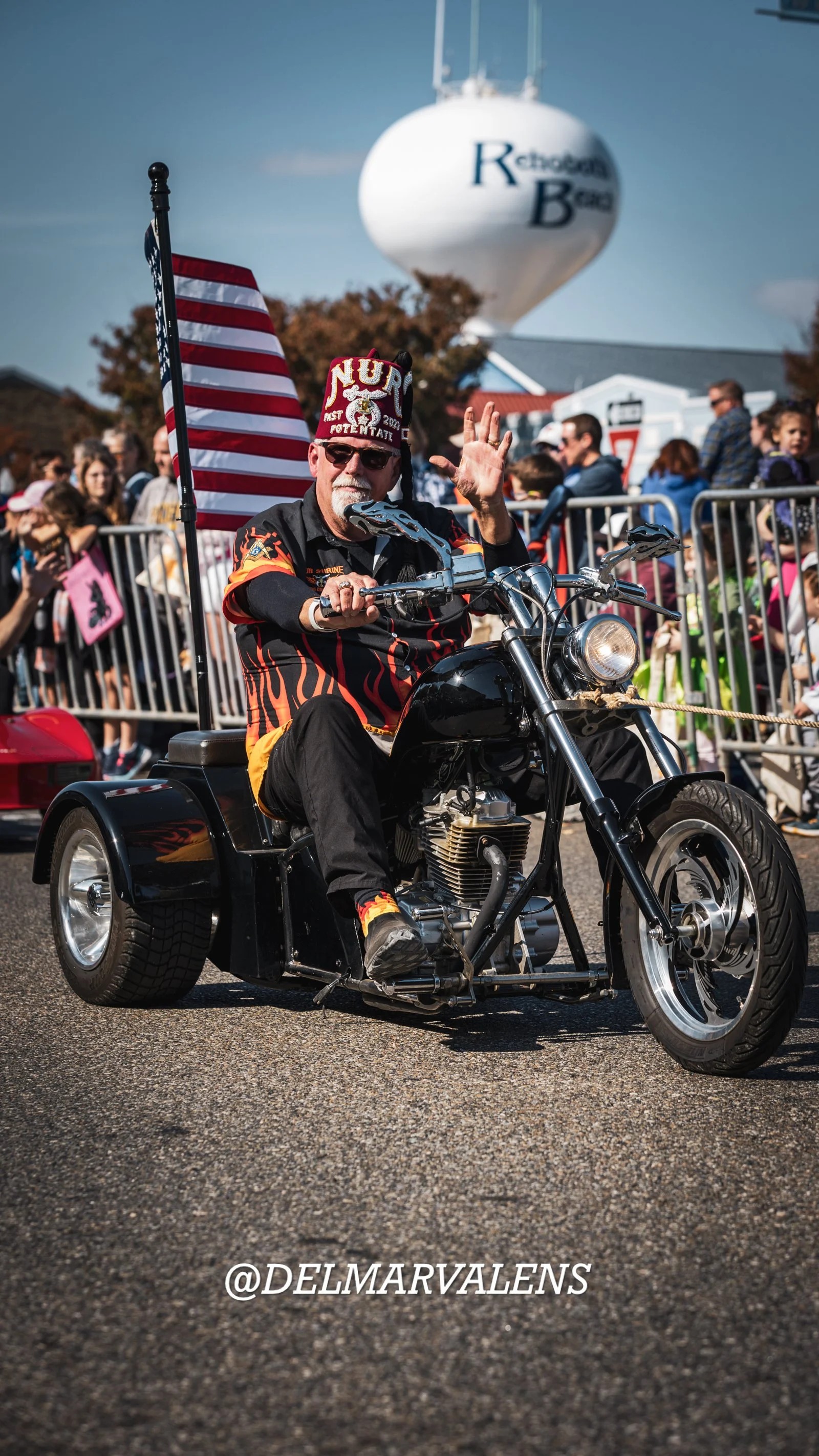 A man riding a motorcycle drawn in a parade, with people watching behind barriers, American flag, and a water tower in the background.