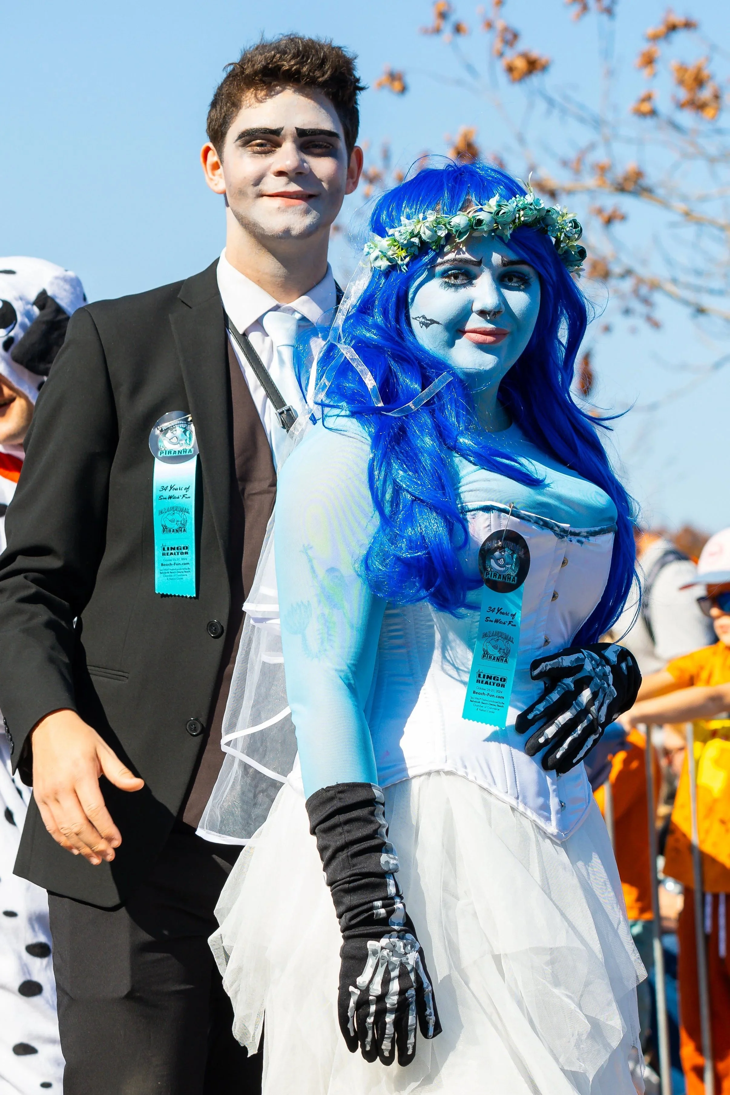 People dressed in costumes at a parade, with a man in a black suit and a woman in a blue wig and white dress, celebrating outdoors.