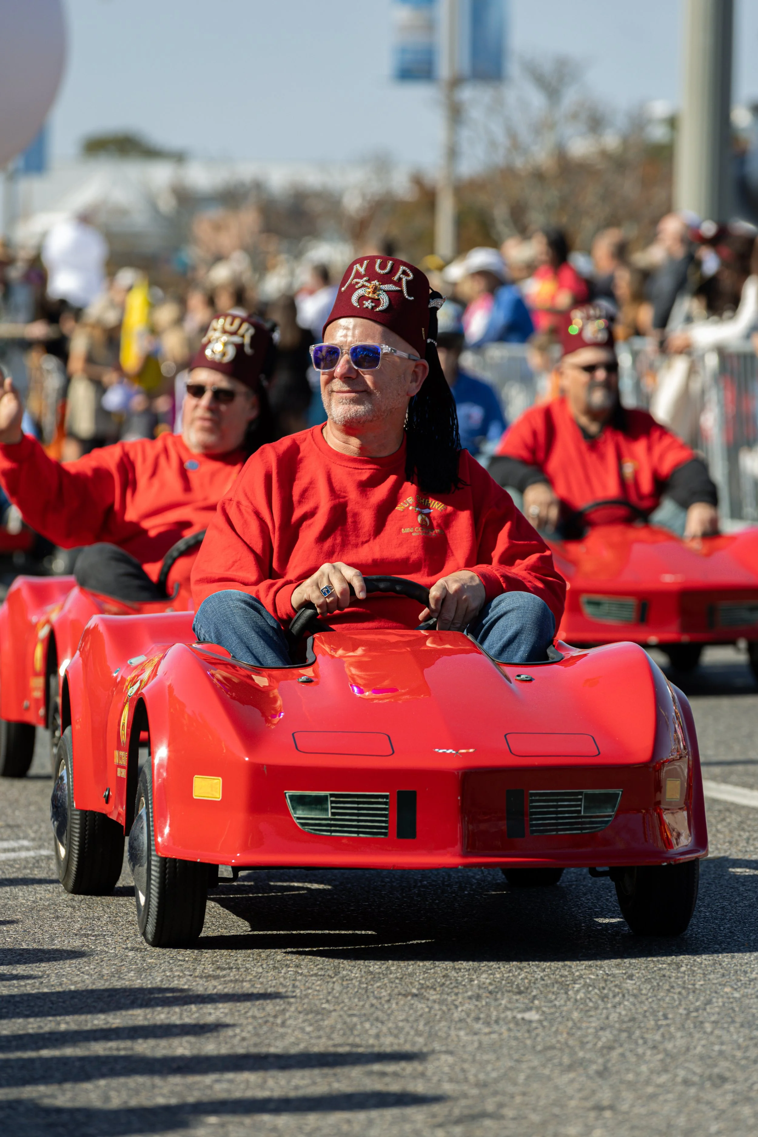 Men dressed in red shirts and wearing red hats with the words 'VET' and an emblem, riding in small, red, toy-like race cars during a parade or event.
