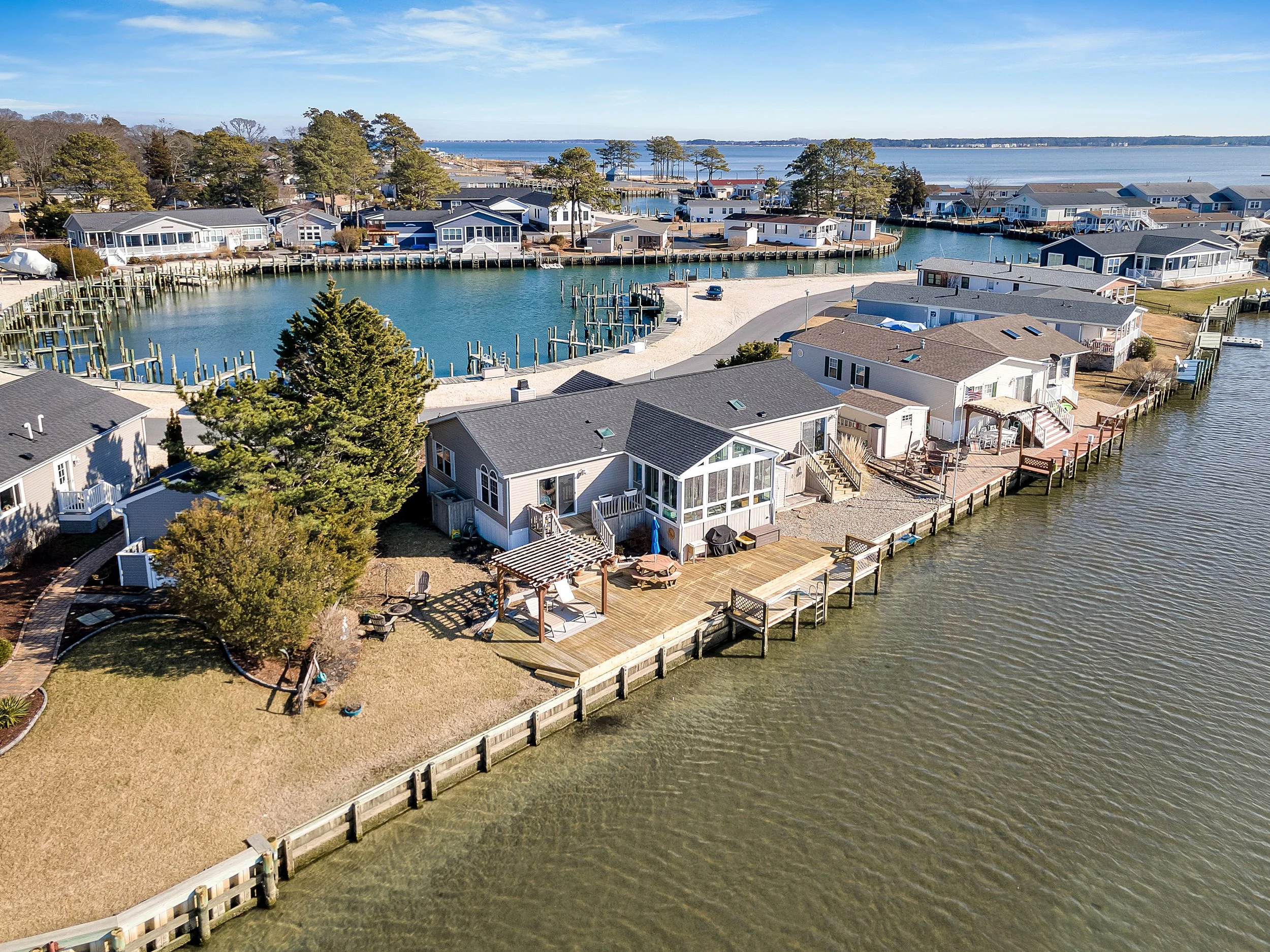 Aerial view of waterfront houses with docks on a bay, showing boats, grassy yards, and trees, with a larger body of water in the background.