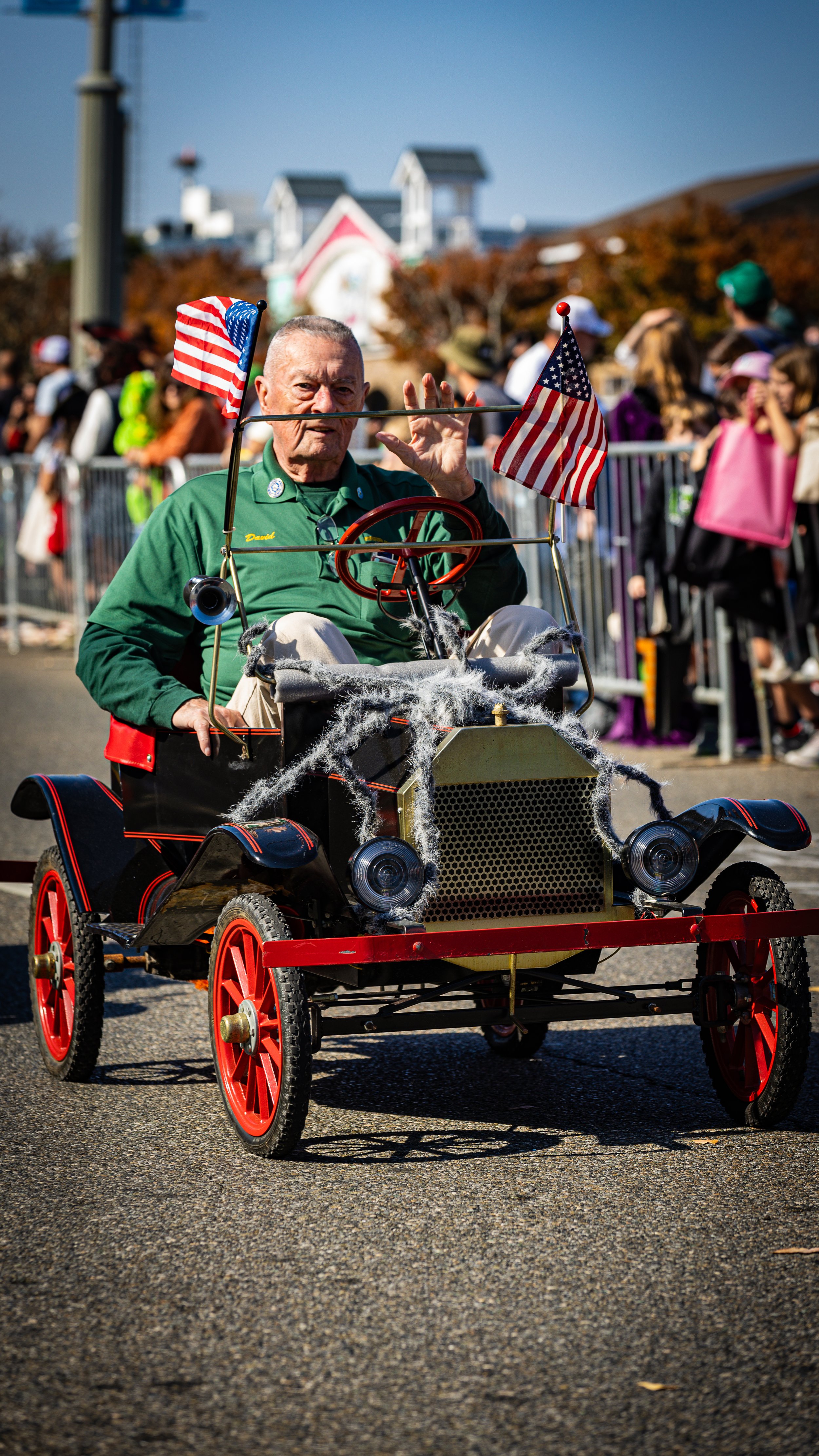A man riding a vintage toy car decorated with cobwebs, American flags, and a red steering wheel, waving during a parade with a crowd of onlookers in the background.