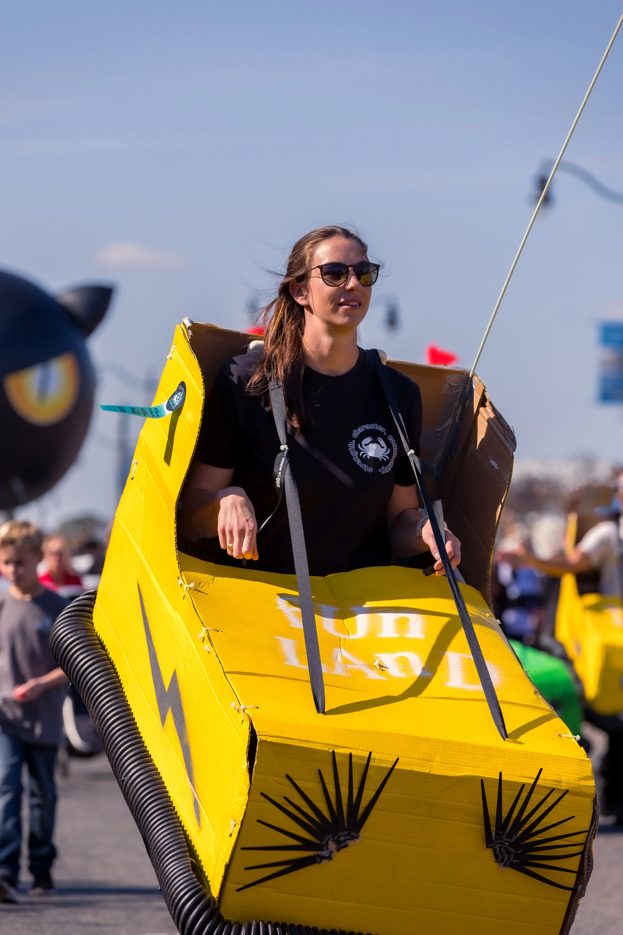 A woman wearing sunglasses and a black T-shirt sits inside a homemade yellow cardboard rocket at an event with people in the background.