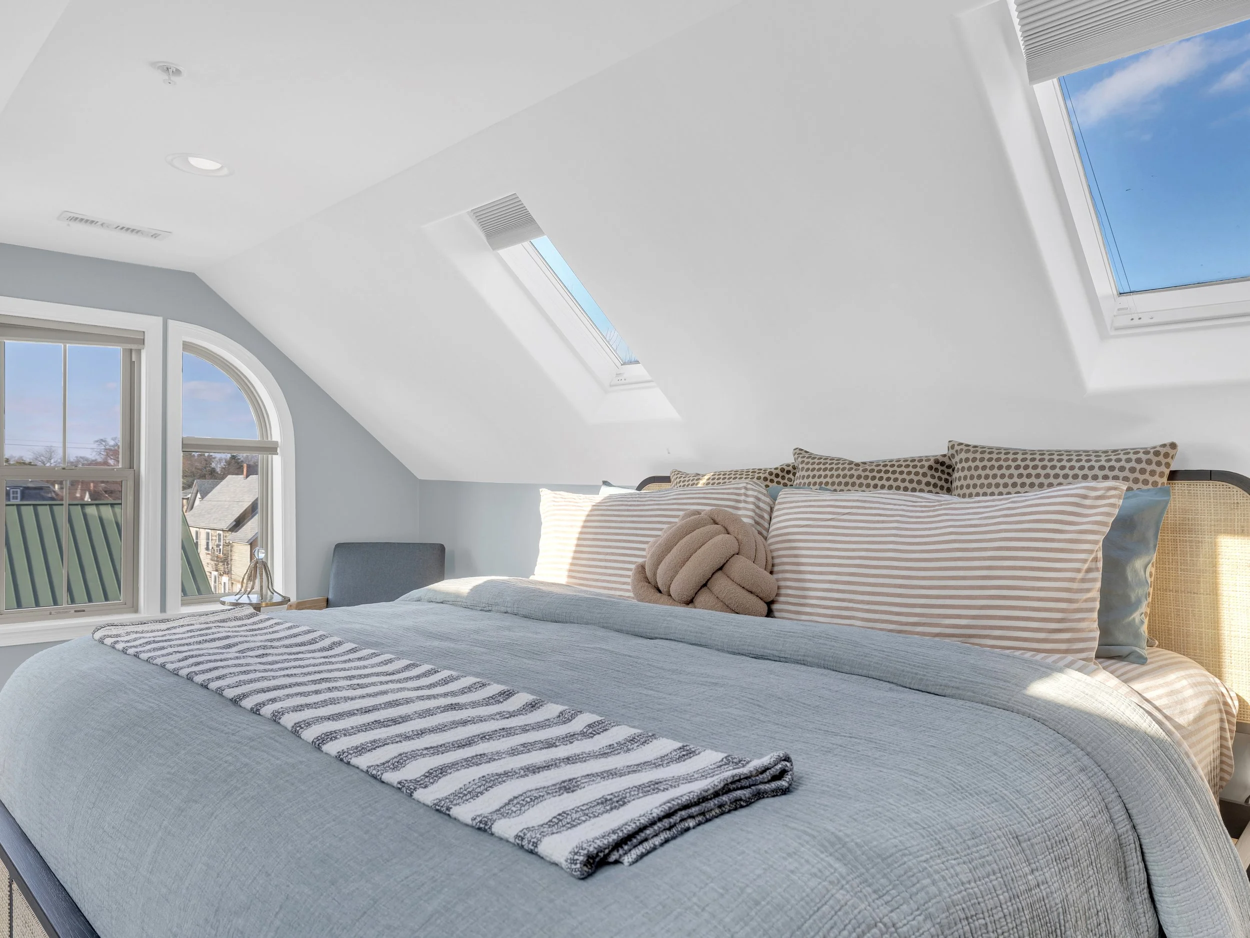 A bedroom with a sloped ceiling and skylights, a bed with various pillows and a striped blanket, and a window showing a neighborhood outside.