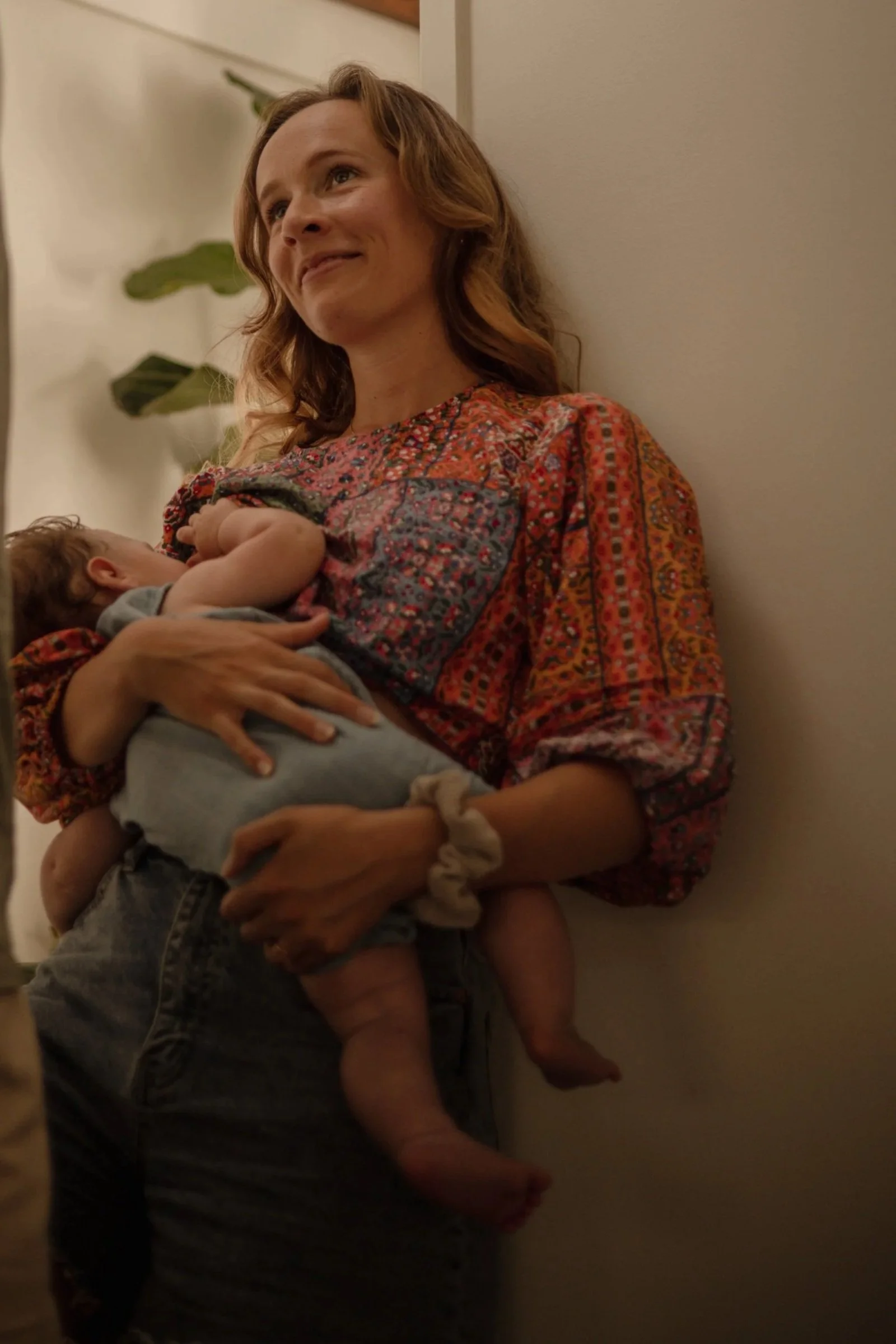A woman is sitting on the floor, leaning against a wall, holding a sleeping baby in her arms, with a contented expression on her face.