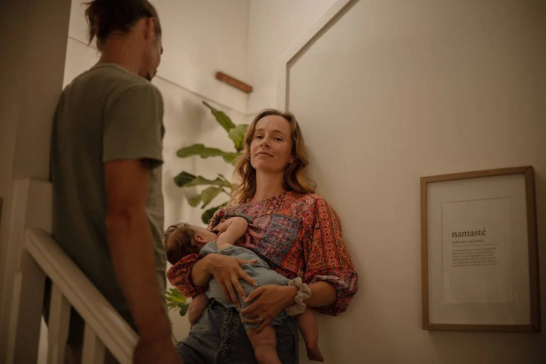Woman holding a sleeping baby in a home hallway, looking confidently at a man standing nearby, with a framed art print on the wall and a large plant in the background.
