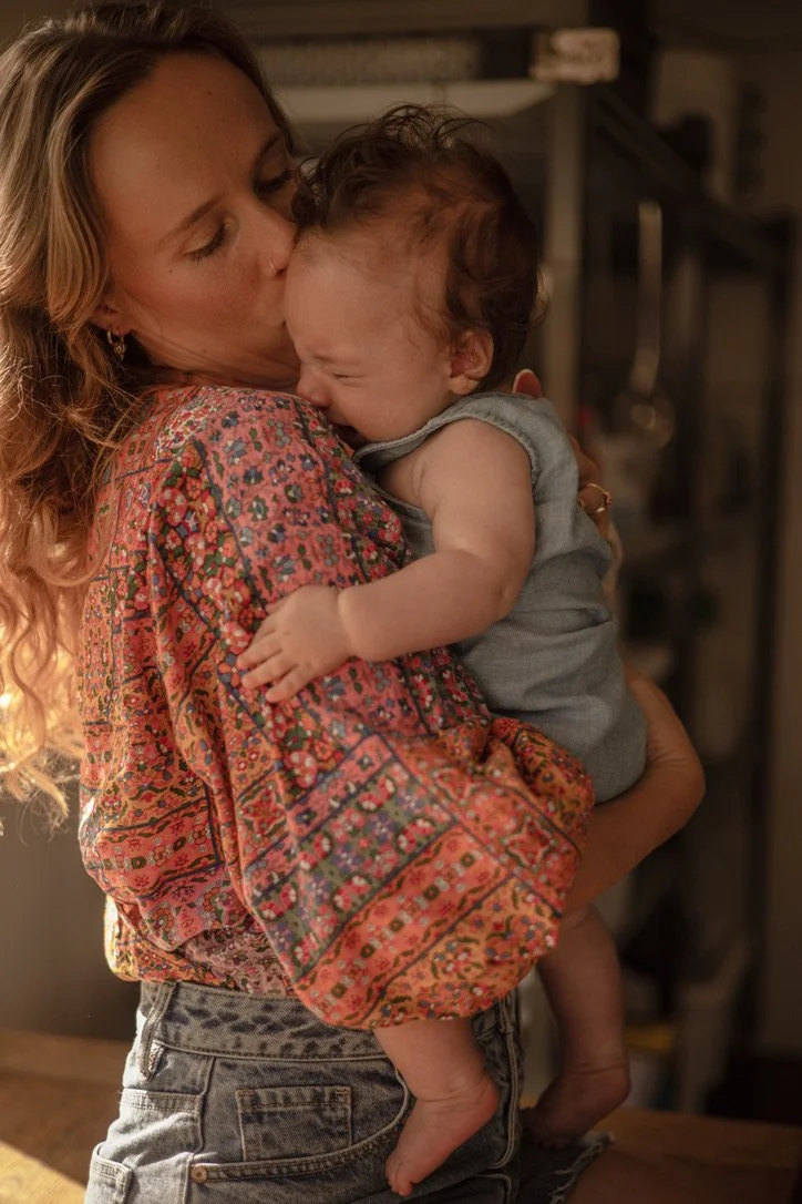 A woman with long, wavy hair holding a crying toddler in her arms, indoors with warm lighting.