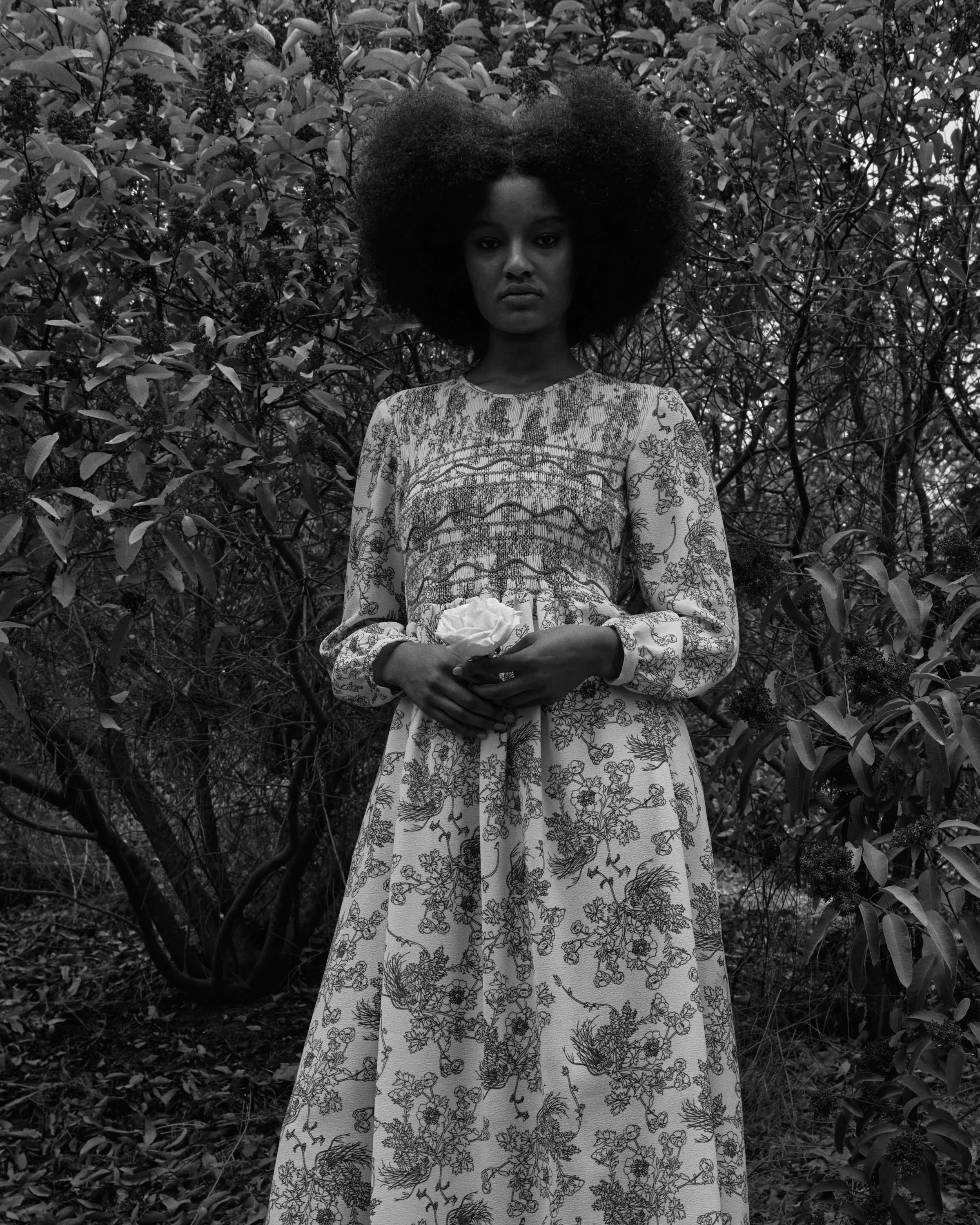 A woman standing outdoors in a garden with dense foliage holding a white flower and a small object, wearing a long patterned dress.