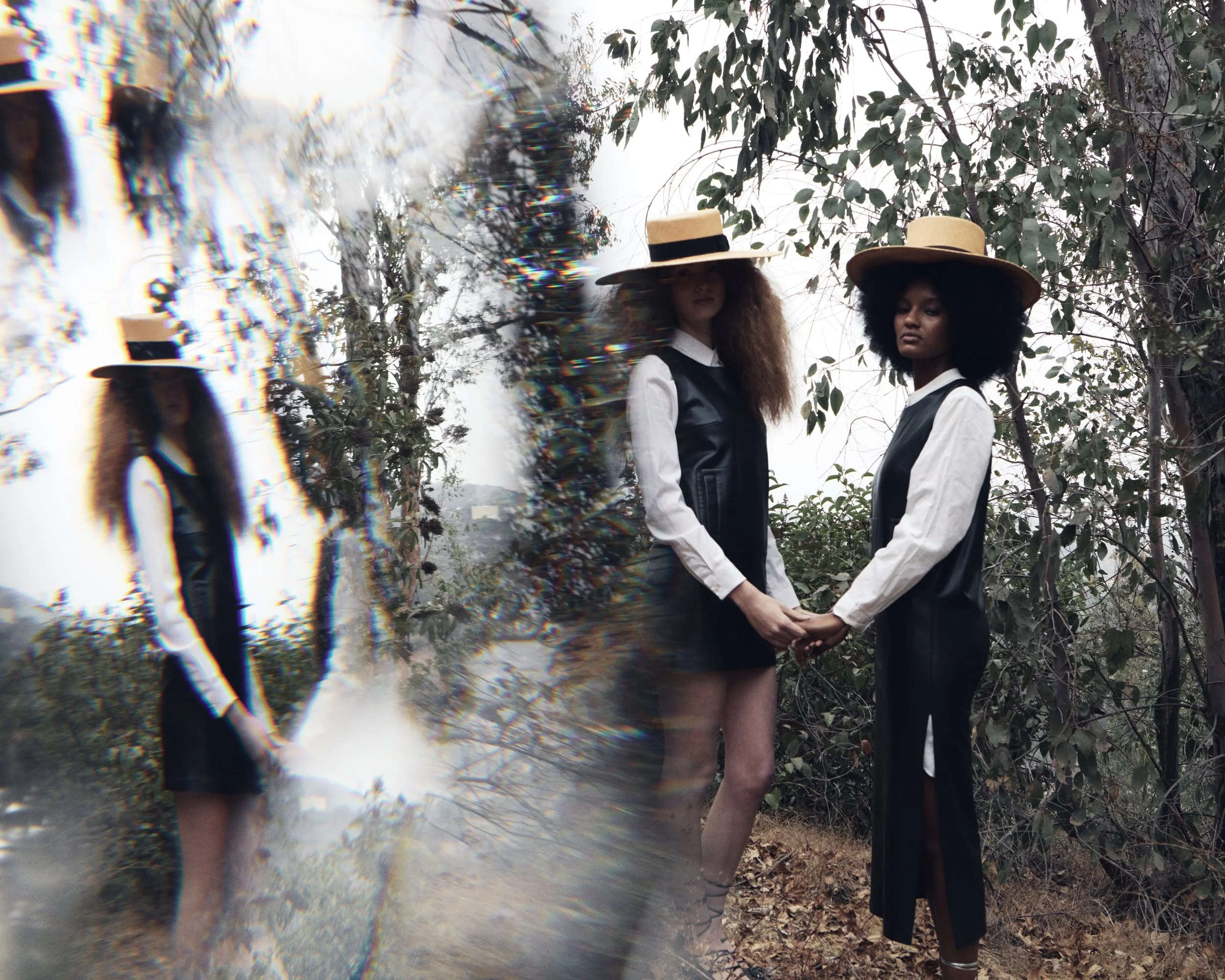 Two women holding hands in a wooded outdoor area, wearing black and white outfits and wide-brimmed straw hats.