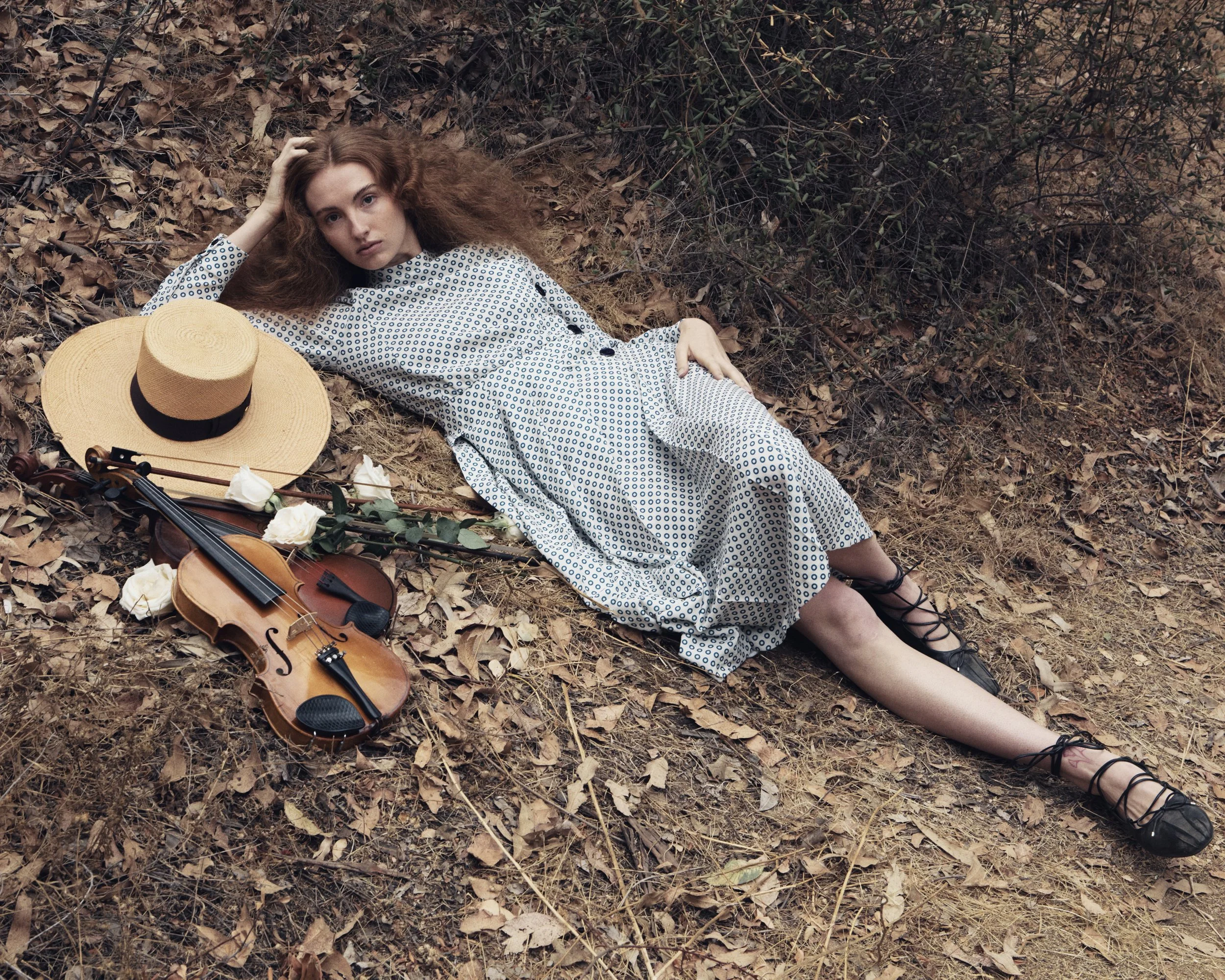 A woman with curly red hair lies on the ground covered with dry leaves and twigs, next to a violin, a white rose, and a wide-brimmed straw hat. She is wearing a long, patterned dress with lace-up black shoes, and looking at the camera with a serious expression.