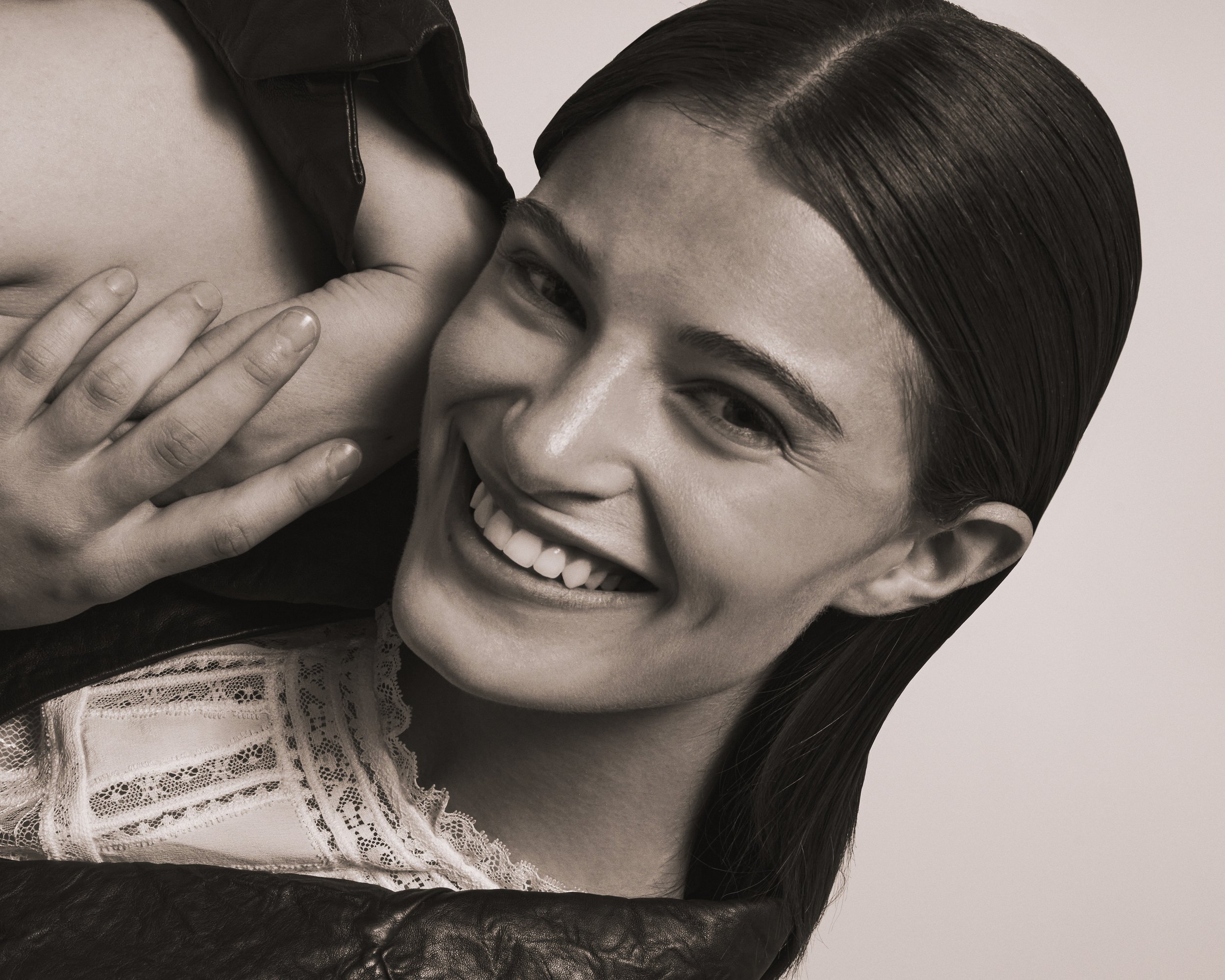 A woman with wet, dark hair smiling while holding a person's shoulder, wearing a lace top and a leather jacket.