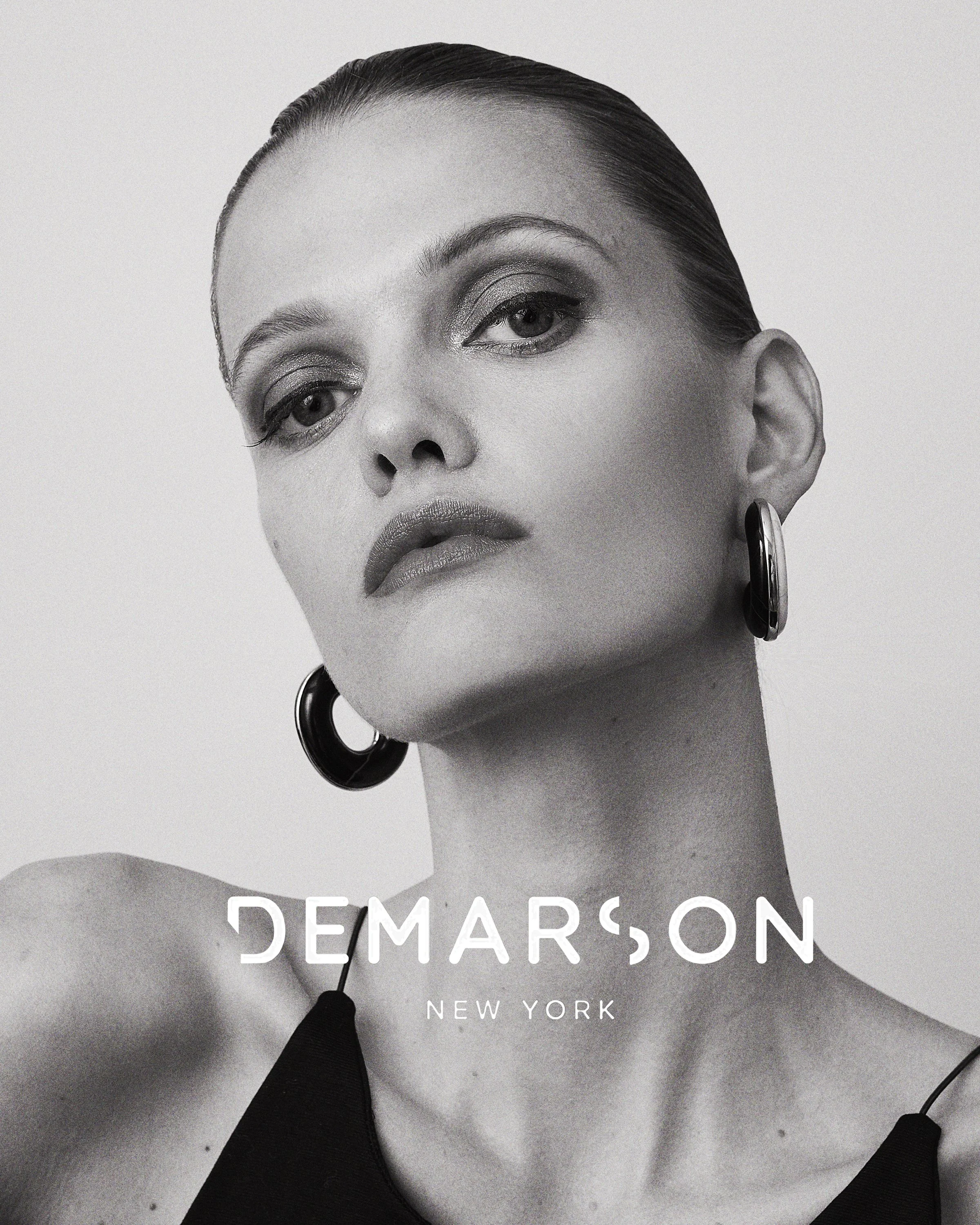 Black and white advertisement for Demarson New York featuring a close-up portrait of a woman with slicked-back hair, large hoop earrings, and bold makeup, with the brand name and location overlaid.