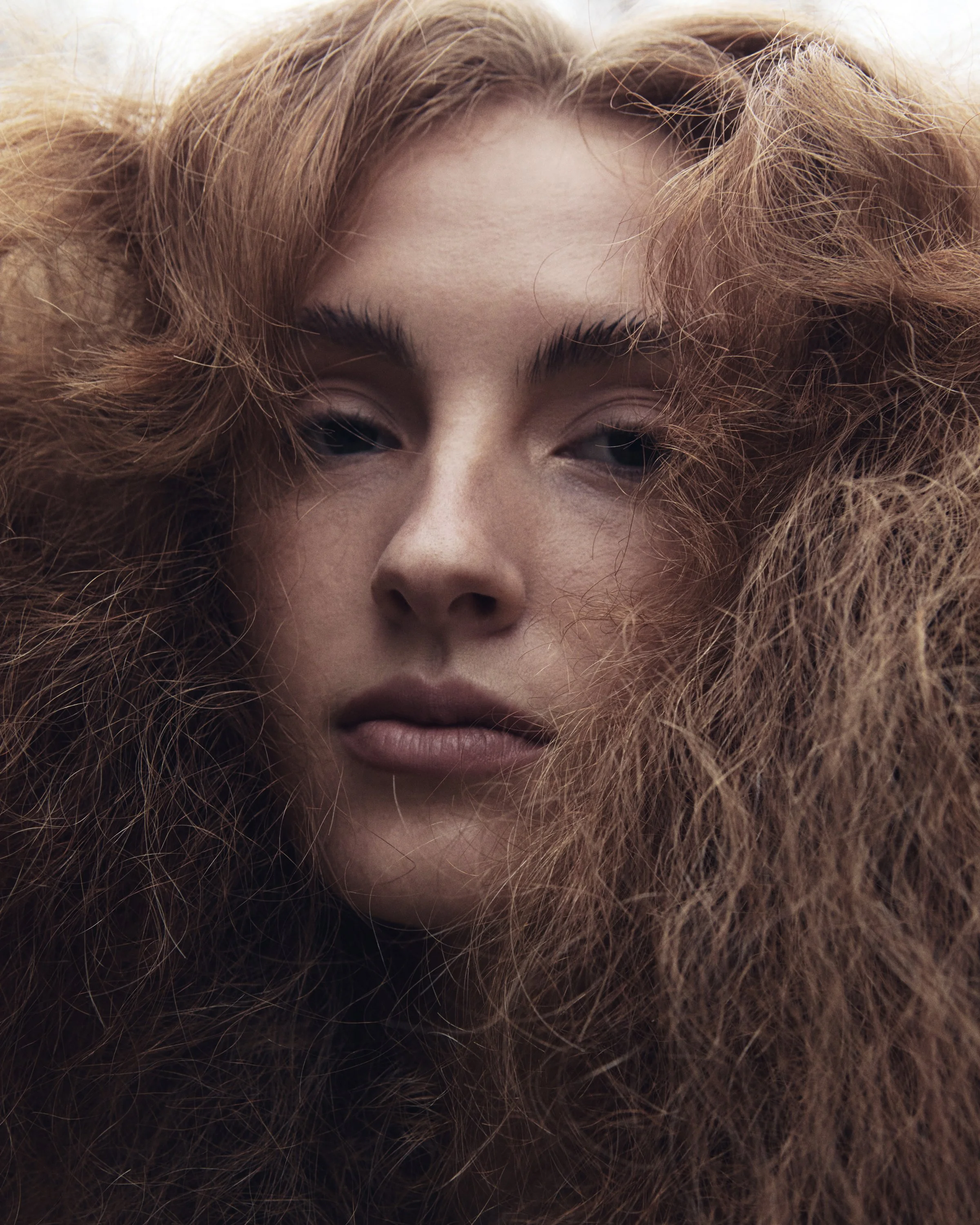 Close-up of a woman with long, curly red hair framing her face, with neutral facial expression and slightly parted lips.