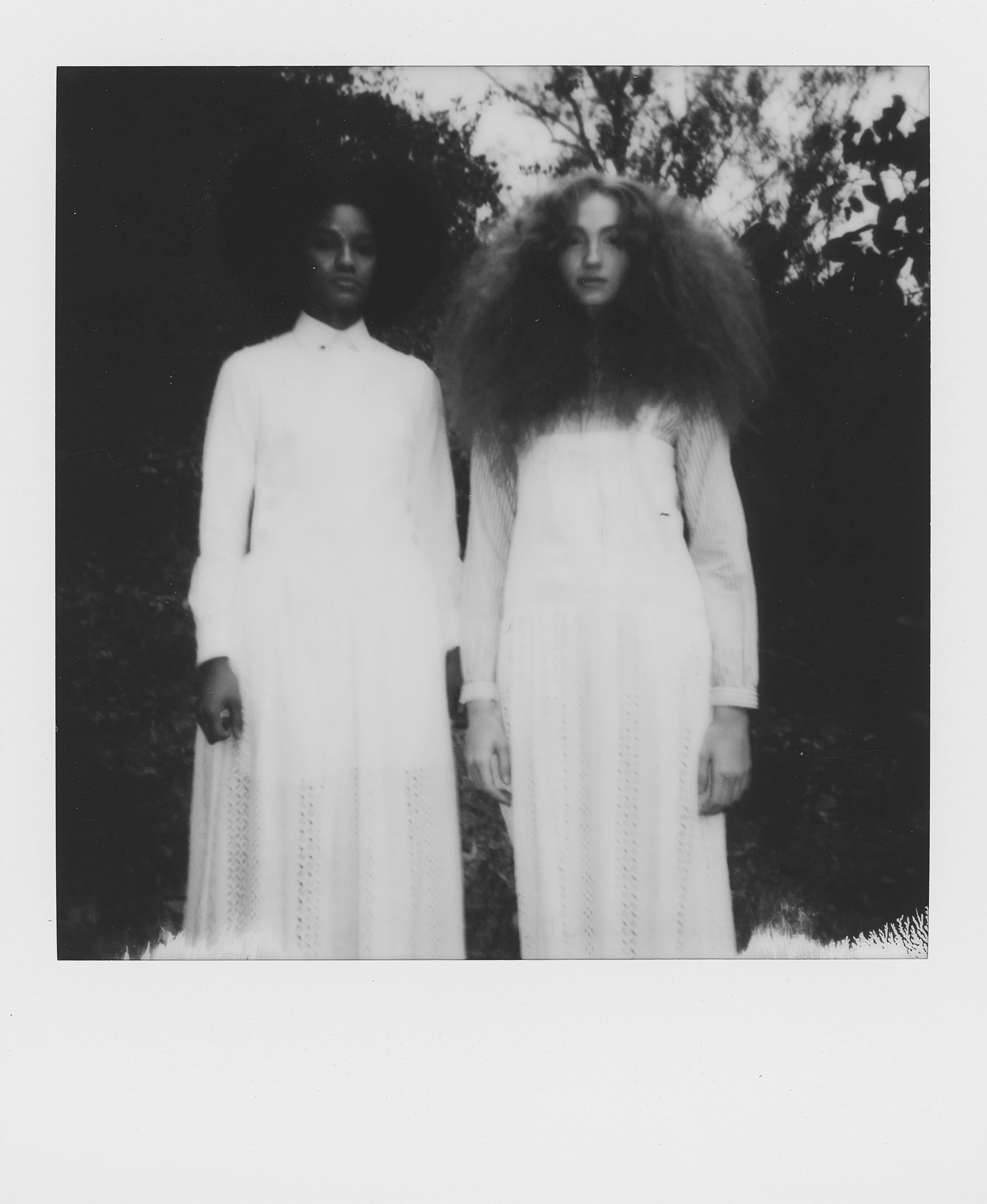 Two women standing outdoors in vintage long white dresses, with one having dark hair and the other with voluminous curly hair.