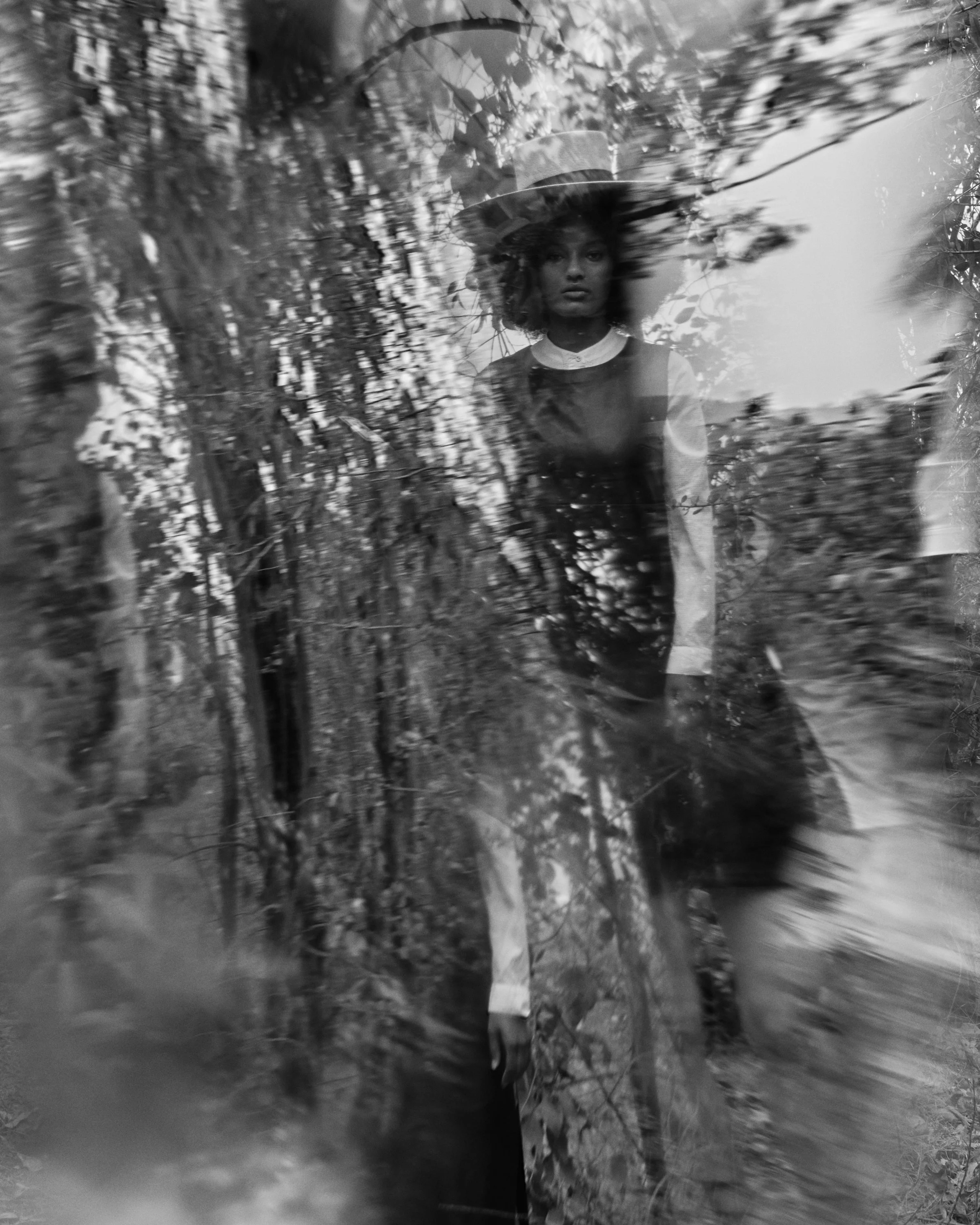 A black and white photograph of a woman standing outdoors among trees, with reflections or double exposure creating a layered effect.