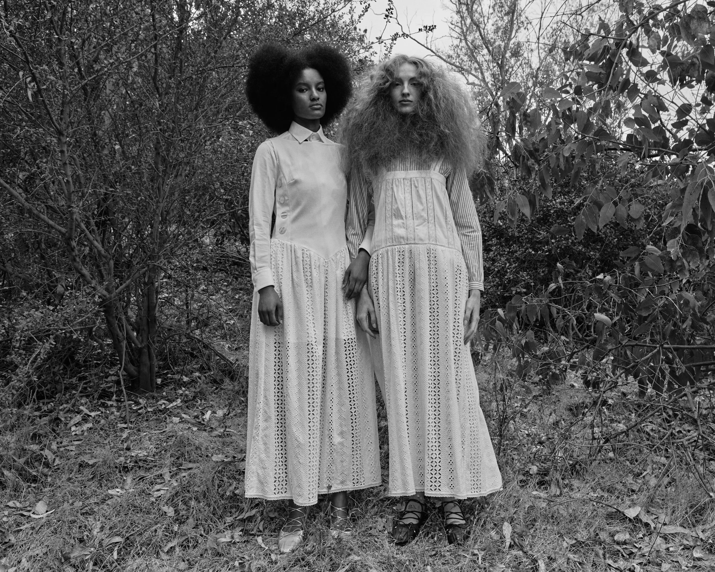 Two women with large, curly hair standing outdoors among trees and bushes, wearing long lace skirts and collared shirts in a black-and-white photo.