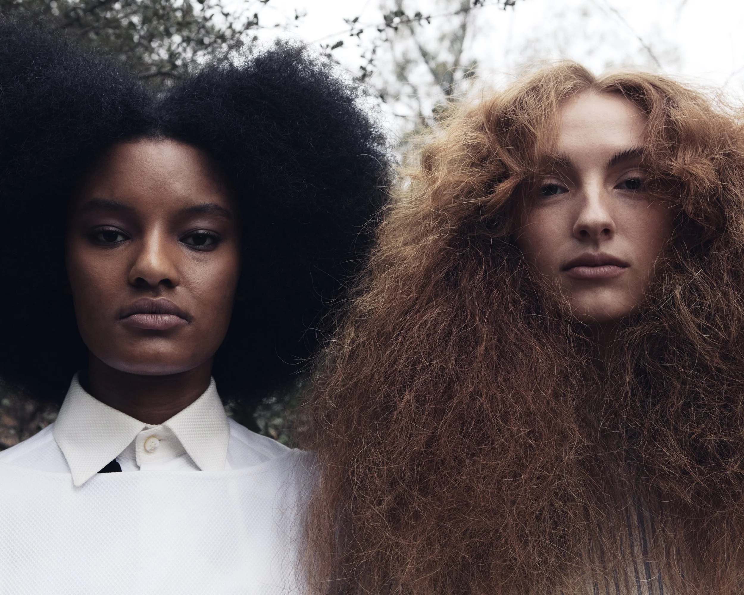 Portrait of two women with voluminous hair, one with black curly hair and the other with red curly hair, standing outdoors against a background of trees.