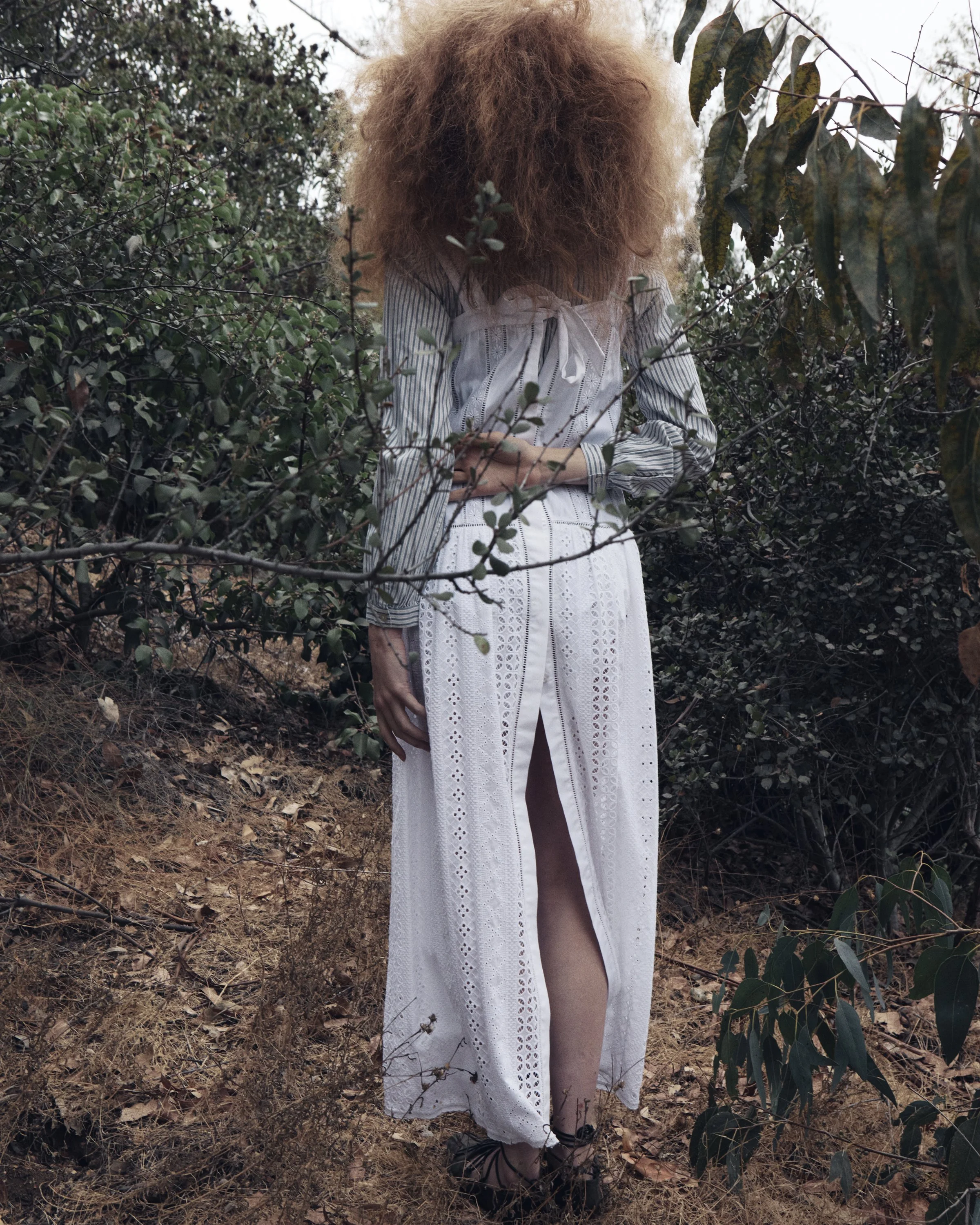 Person with voluminous curly red hair wearing a long white eyelet dress with a slit, standing among bushes in a natural outdoor setting.