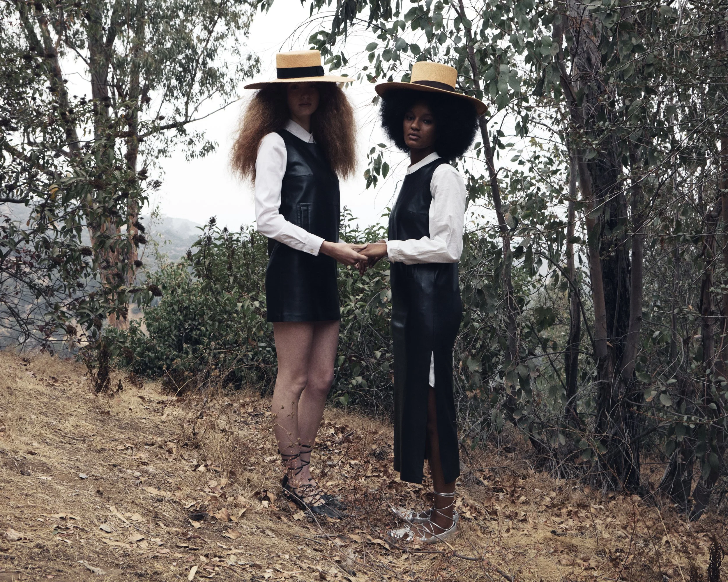 Two women standing outdoors on a dirt path in a wooded area, holding hands and looking at the camera. They are wearing black dresses with white shirts underneath, wide-brimmed straw hats, and high-heeled shoes.