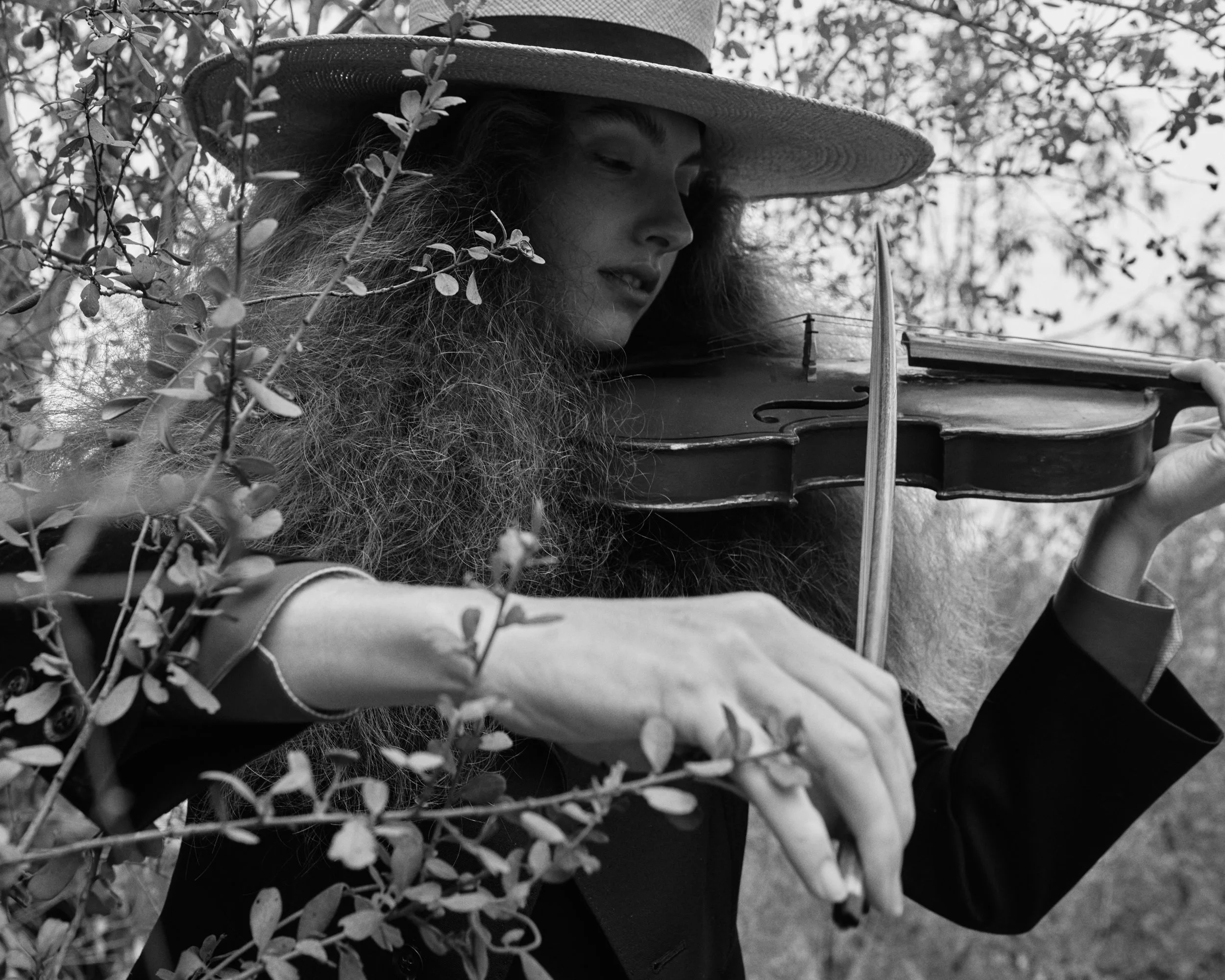 A woman with curly hair wearing a wide-brimmed hat playing the violin outdoors among branches and leaves.