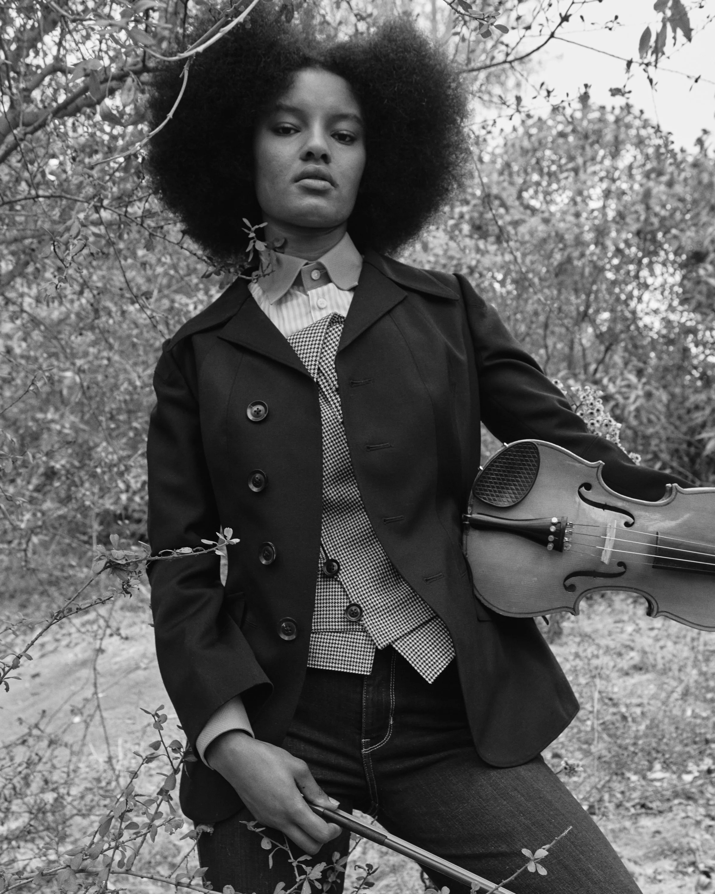 A woman with an afro hairstyle holding a violin and a bow, standing outdoors among trees and bushes, posing confidently in a stylish outfit.
