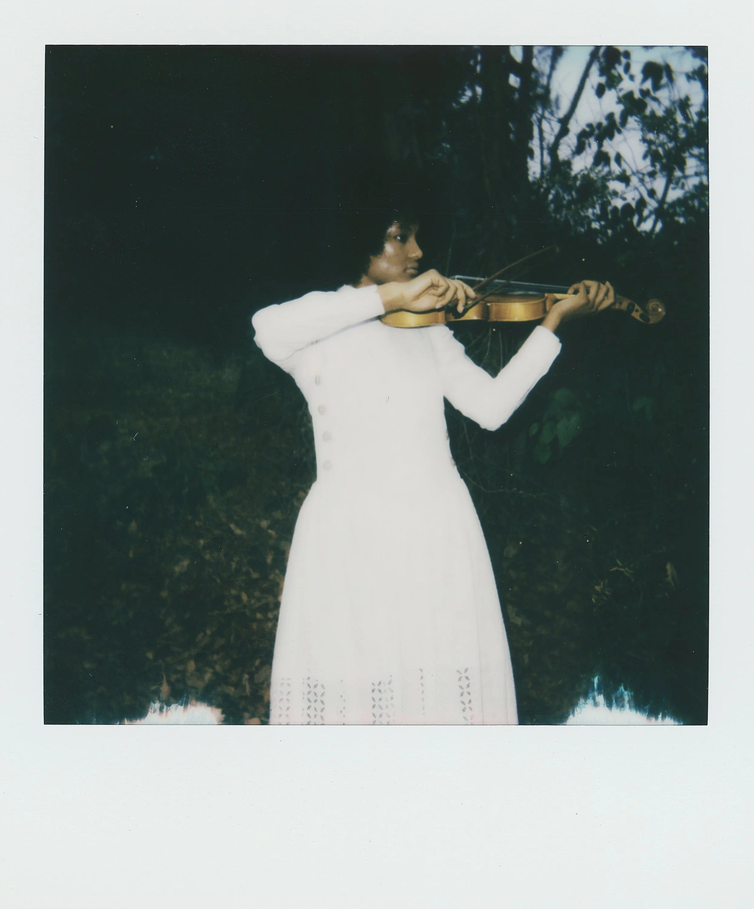 A woman in a white dress playing the violin outdoors, with trees and foliage in the background.