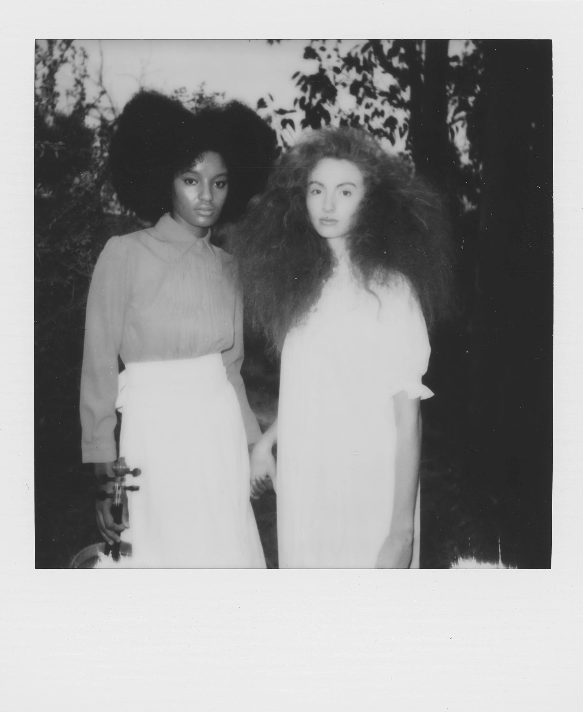 Two women with large, voluminous hair standing outdoors, holding hands, black and white photo.