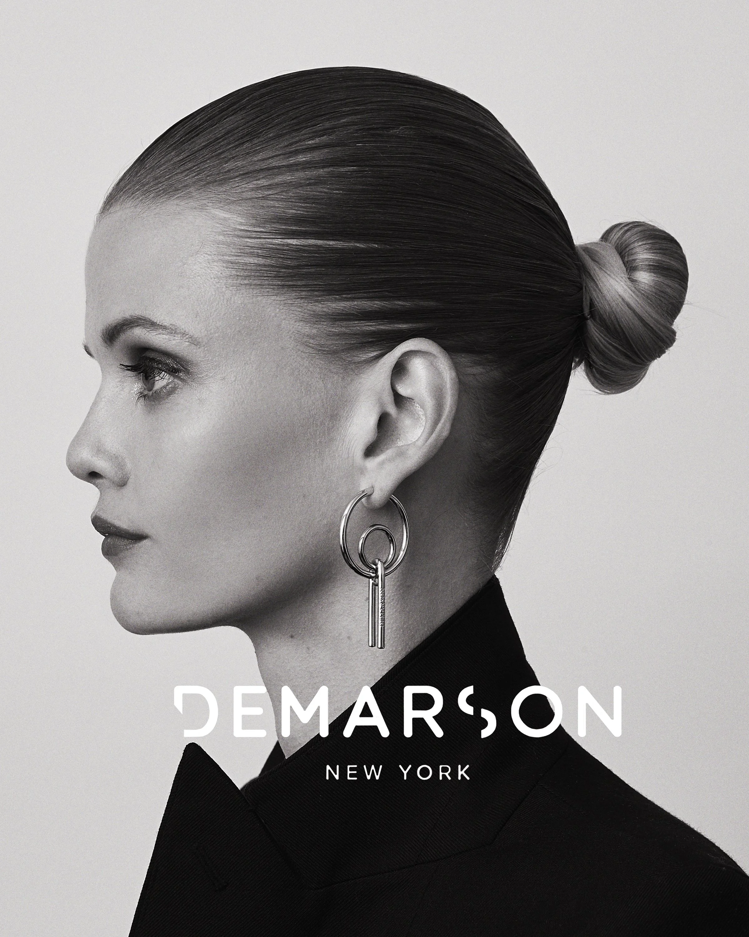 Black and white profile of a woman with sleek hair in a bun, wearing geometric earrings and a dark blazer, with the text 'Dемарсон New York' overlaid.