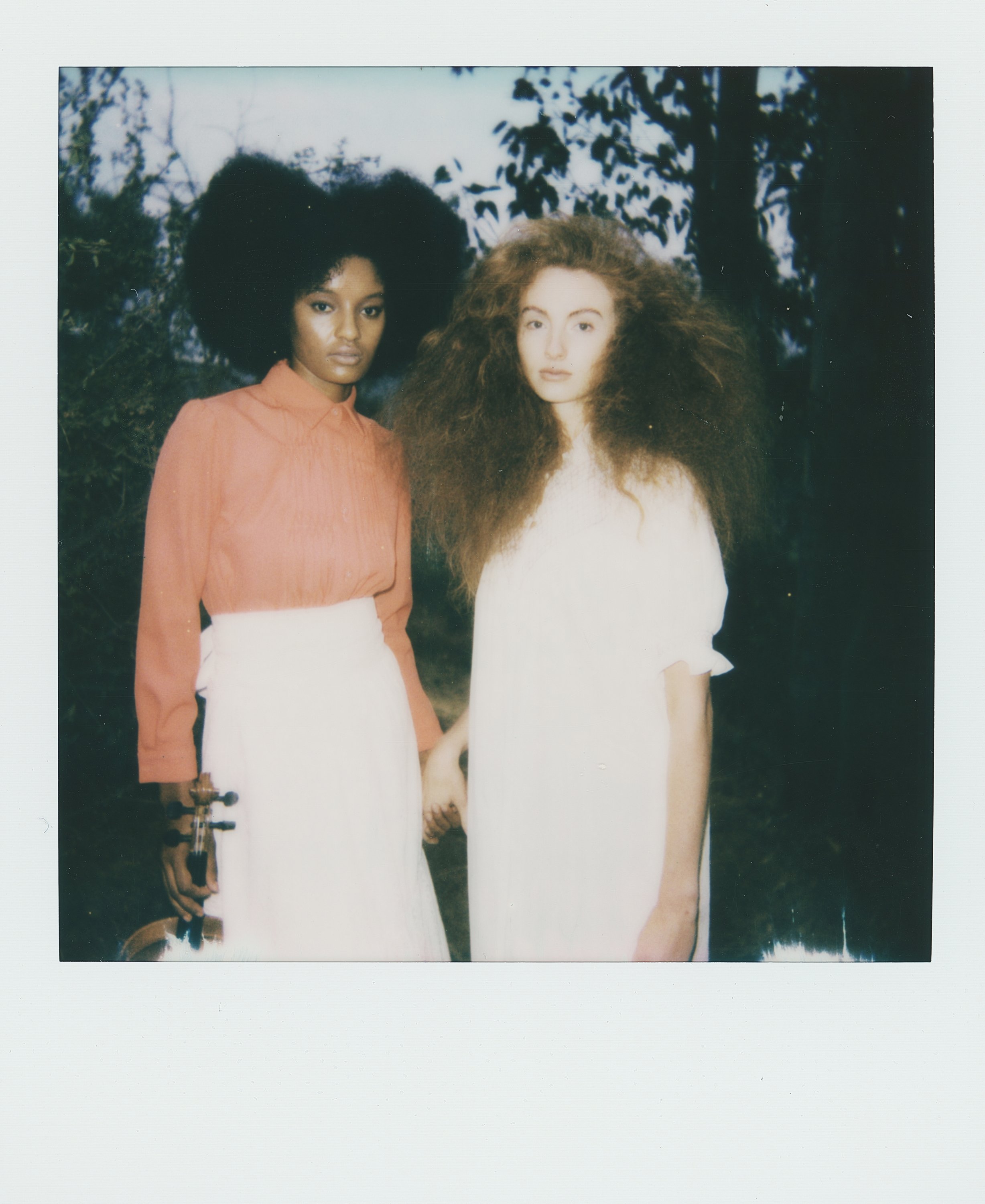 Two women standing outdoors holding hands, one with natural curly black hair wearing an orange blouse and white skirt, the other with voluminous red hair wearing a white dress, with trees in the background.