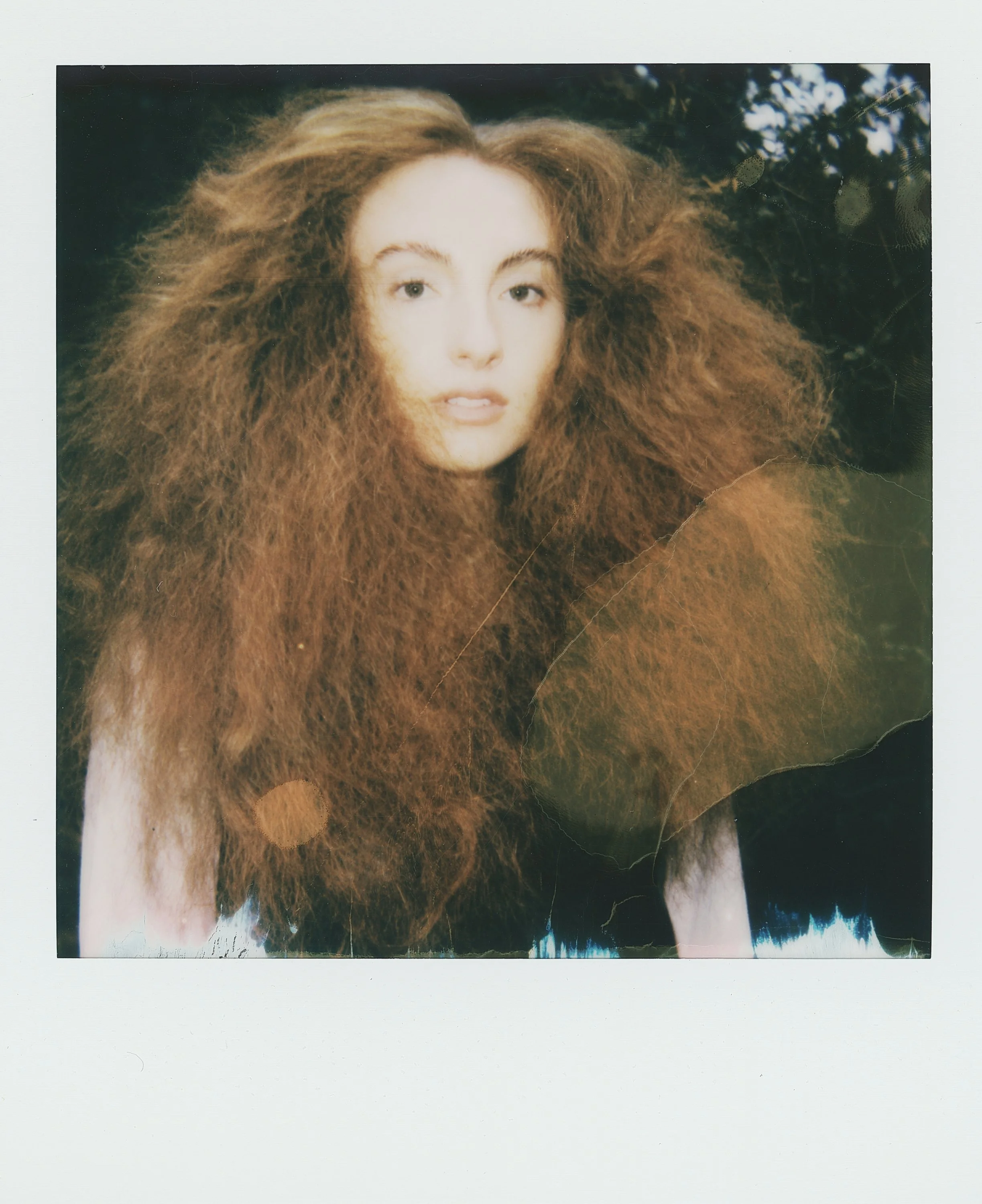 A woman with very long, voluminous, curly red hair outdoors, with a dark background and foliage visible.