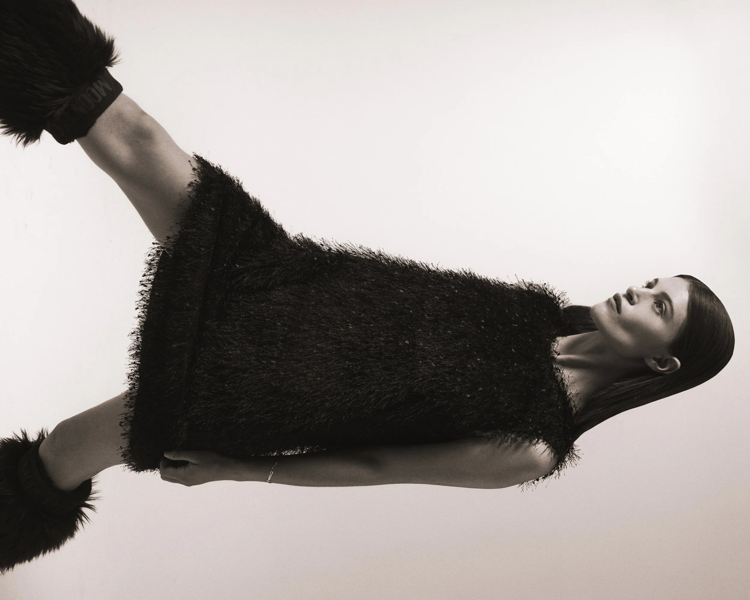A woman in a black, textured, sleeveless dress looking to her left, with long straight hair, standing against a plain white background.