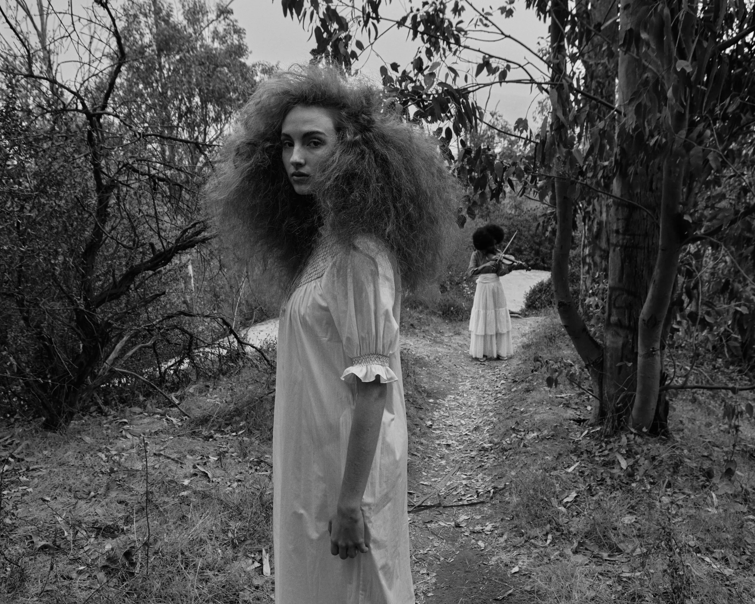 A black-and-white photo of a woman with long, wild hair standing on a dirt path in a wooded area, with a woman playing a violin in the background.
